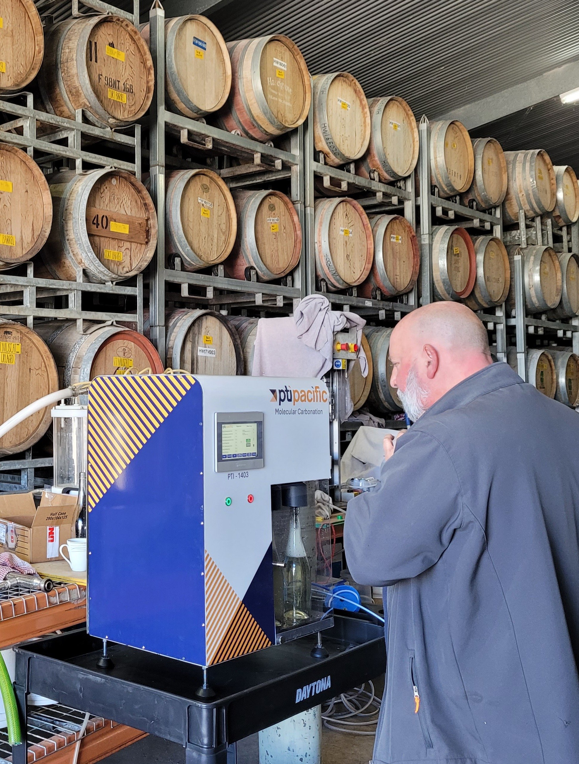 A man operating a molecular carbonation machine in a brewery or laboratory, with barrels stacked on racks in the background.