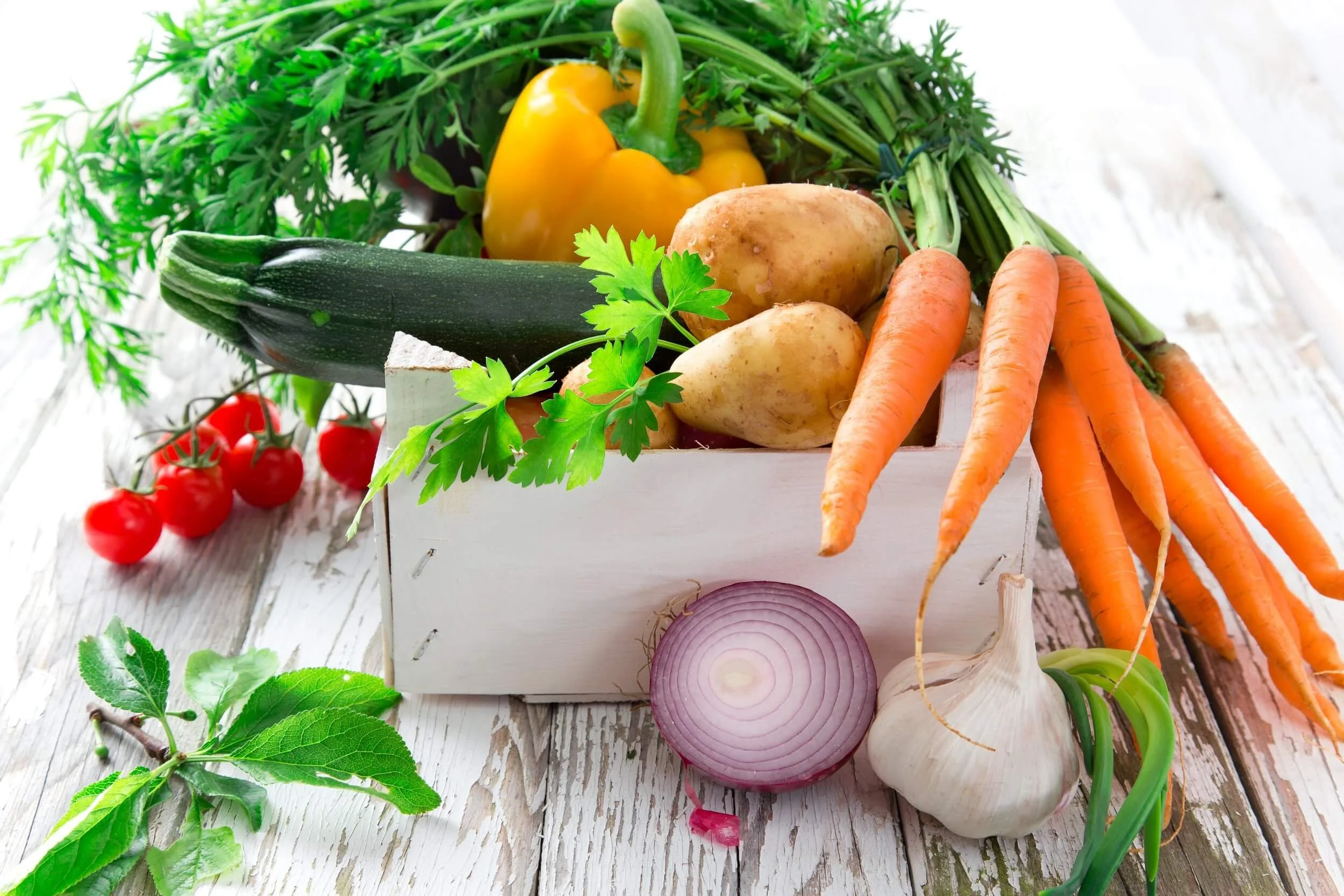 Fresh vegetables including cherry tomatoes, zucchini, carrots, potatoes, red onion, garlic, green herbs, yellow bell pepper, and leafy greens in a white wooden box on a rustic wooden surface.