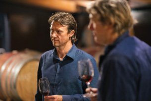 Two men in a wine cellar holding glasses of red wine, with wine barrels in the background.