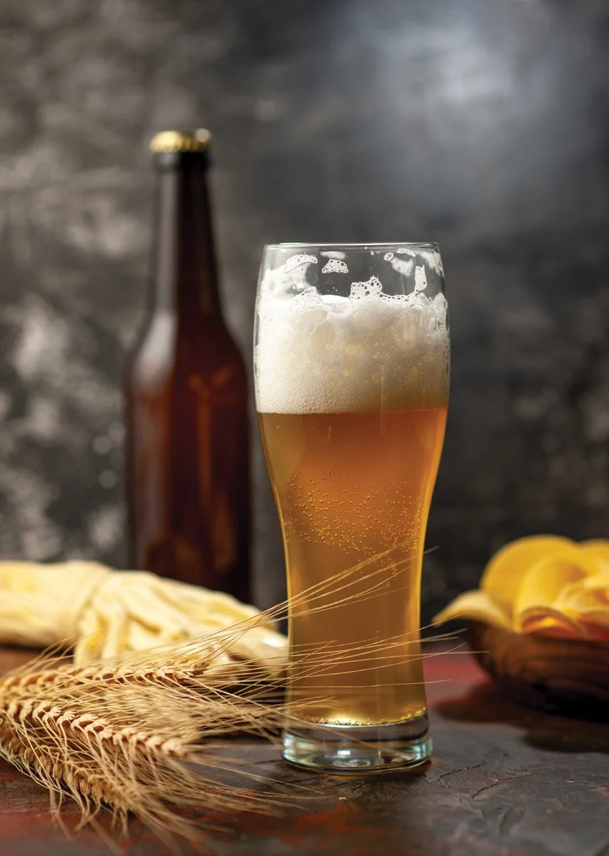 Tall glass of beer with frothy head, a brown beer bottle, wheat stalks, and sliced citrus.