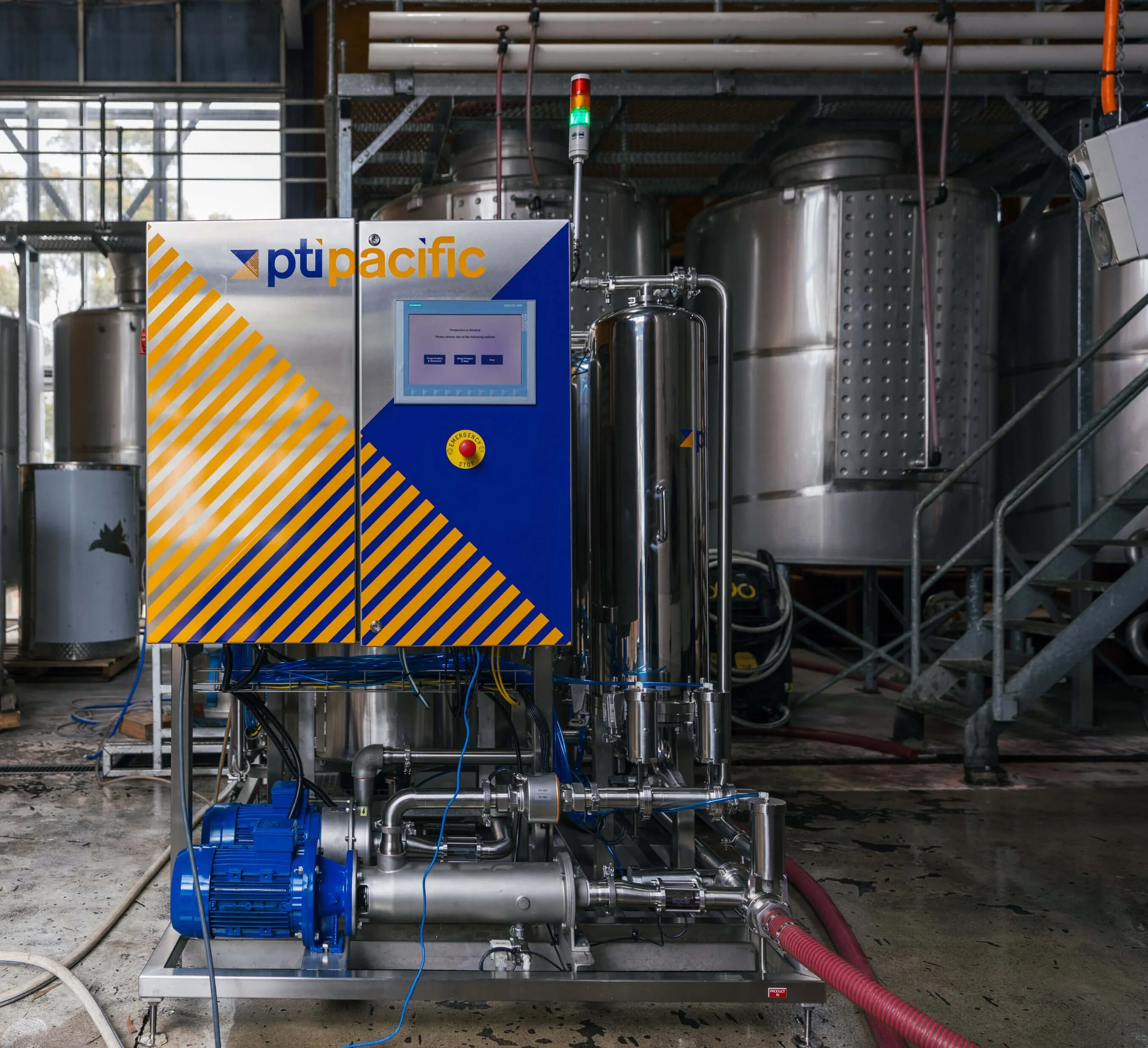 Industrial machinery in a brewery featuring a control panel with a screen and emergency stop button, surrounded by stainless steel tanks and pipes.