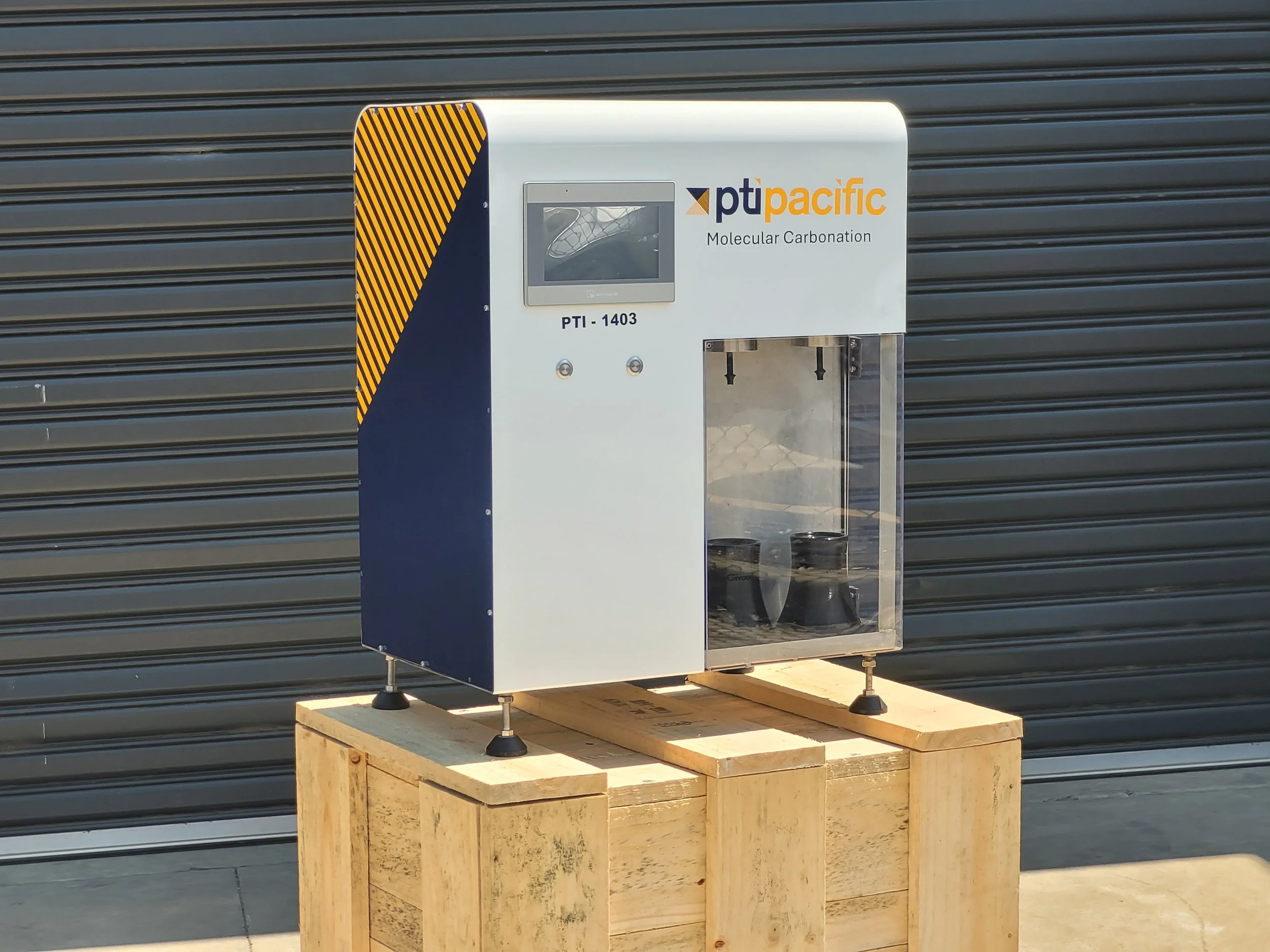 Molecular carbonation machine labeled PtiPacfic on a wooden platform in front of a black metal roll-up door.
