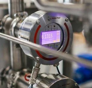 Close-up of an industrial digital pressure sensor with a purple display showing a measurement of 6.1315, attached to piping in a manufacturing setting.