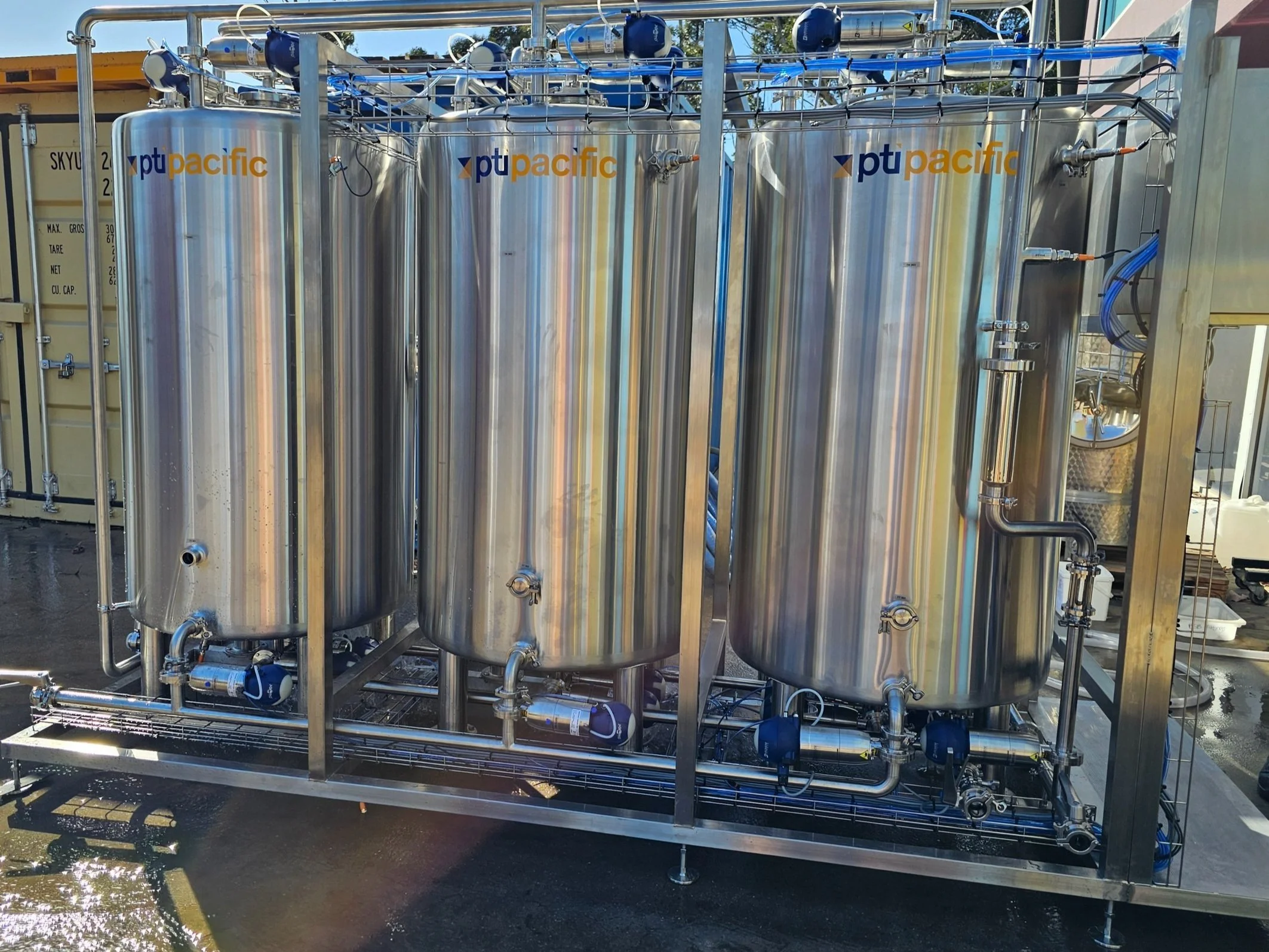 Three large stainless steel tanks with the PT Pacific logo, connected by pipes and valves, outdoors on a metal frame.
