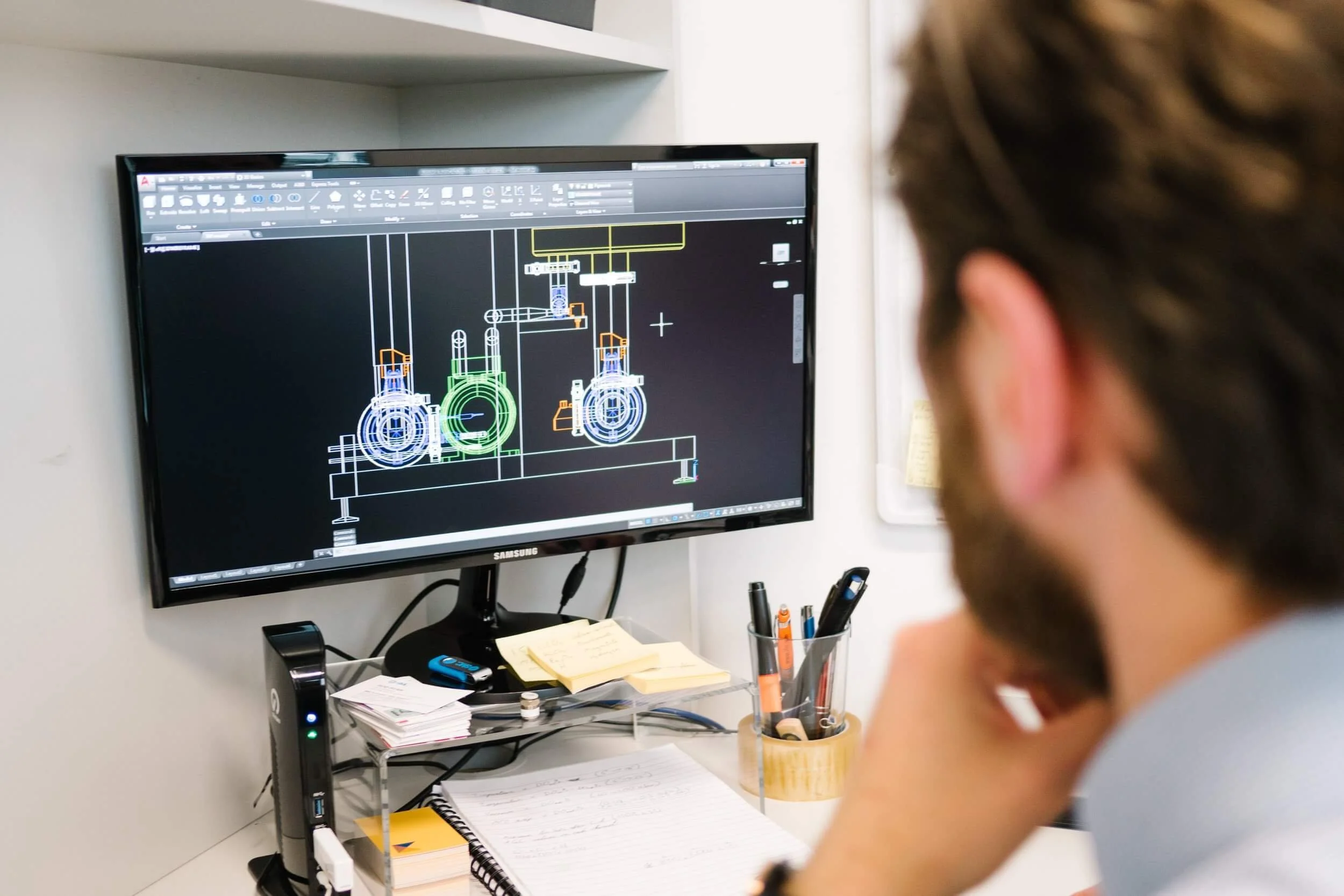 A man with a beard looking at a computer monitor displaying a technical CAD drawing of mechanical parts.