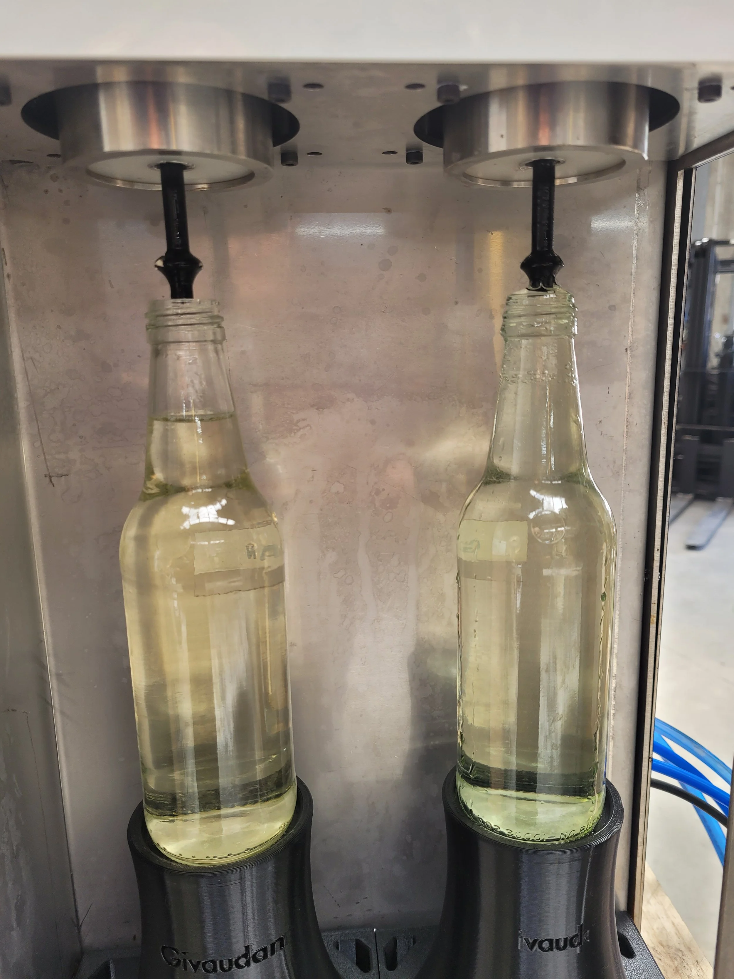 Two glass bottles being filled with a clear liquid at a beverage filling station with black nozzles.