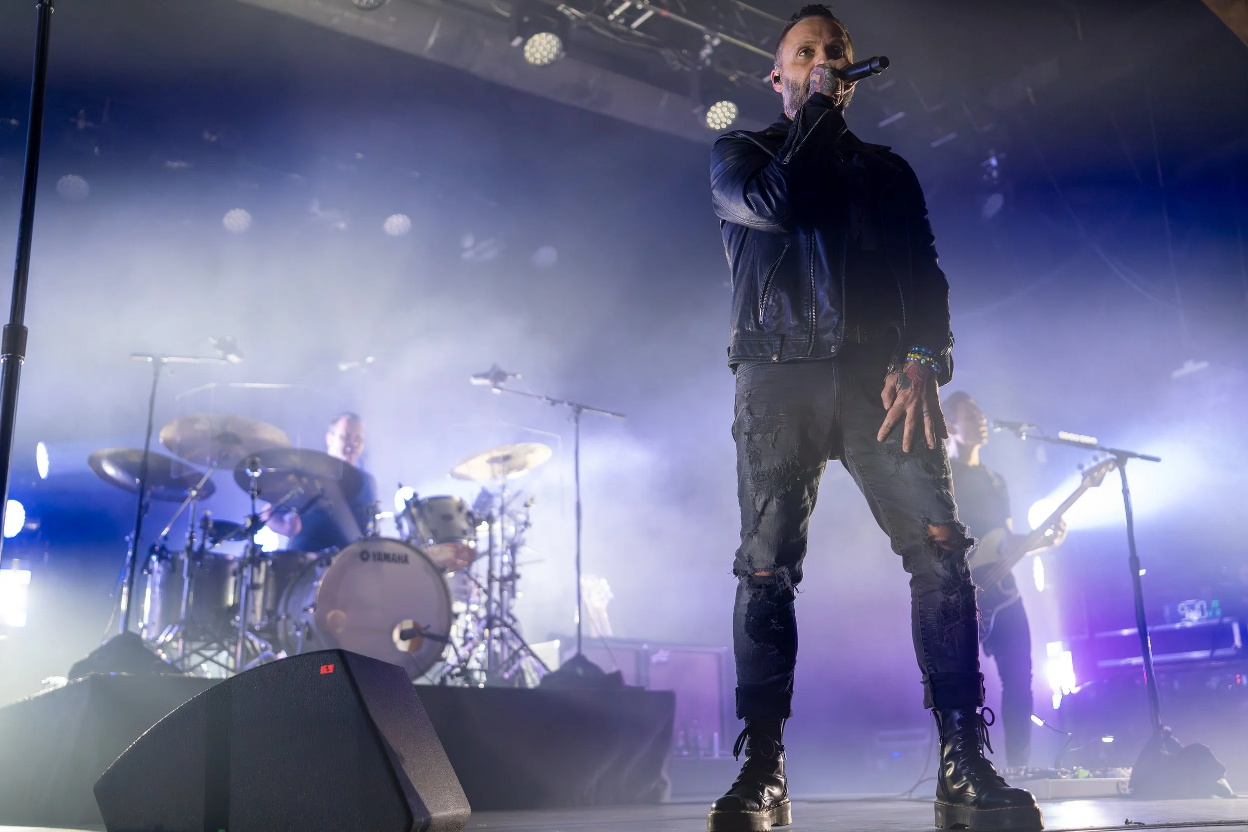 A male singer with tattoos, wearing a black leather jacket and ripped jeans, performing on stage with a microphone, with a drummer and guitarist in the background, stage lights, and fog effects.