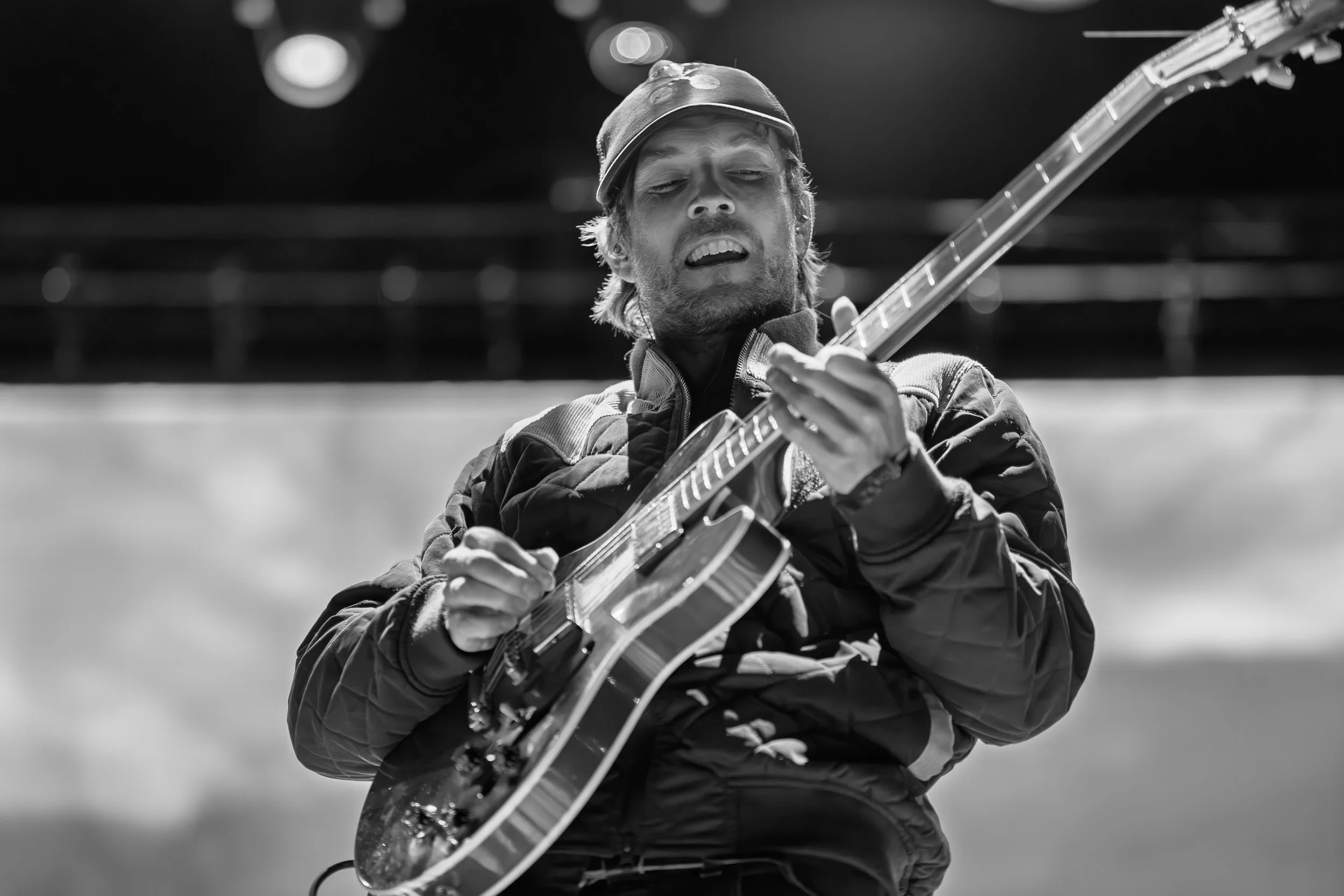 A man playing an acoustic guitar outdoors, wearing a cap and a jacket, with a focused expression.