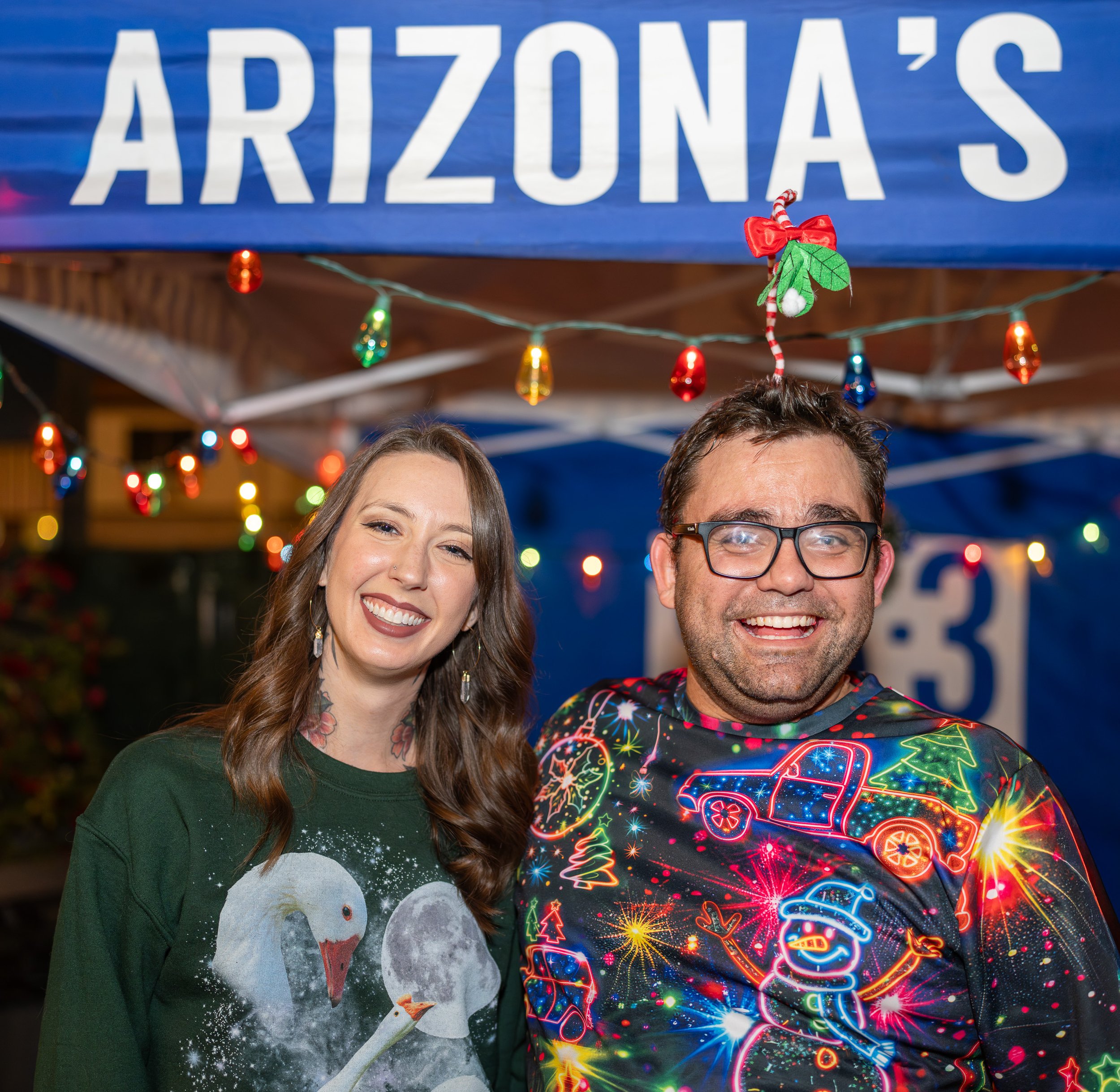 Two smiling people at a festive holiday event, standing under a blue banner with the partial text 'ARIZONA'S.' One person is wearing a Christmas sweater with a swan and moon design, and the other is wearing a black sweatshirt decorated with colorful 