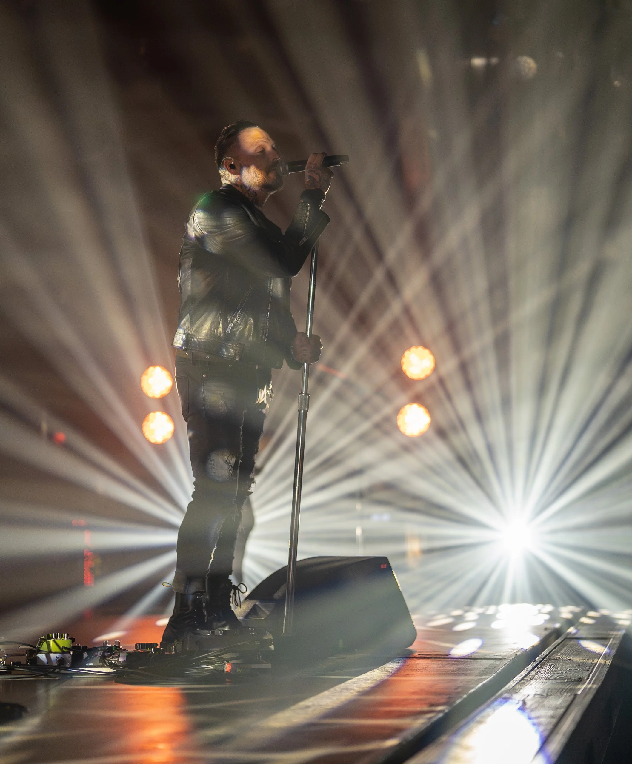 Male singer performing on stage with a microphone, wearing a black leather jacket and ripped jeans, with bright stage lights shining behind him.