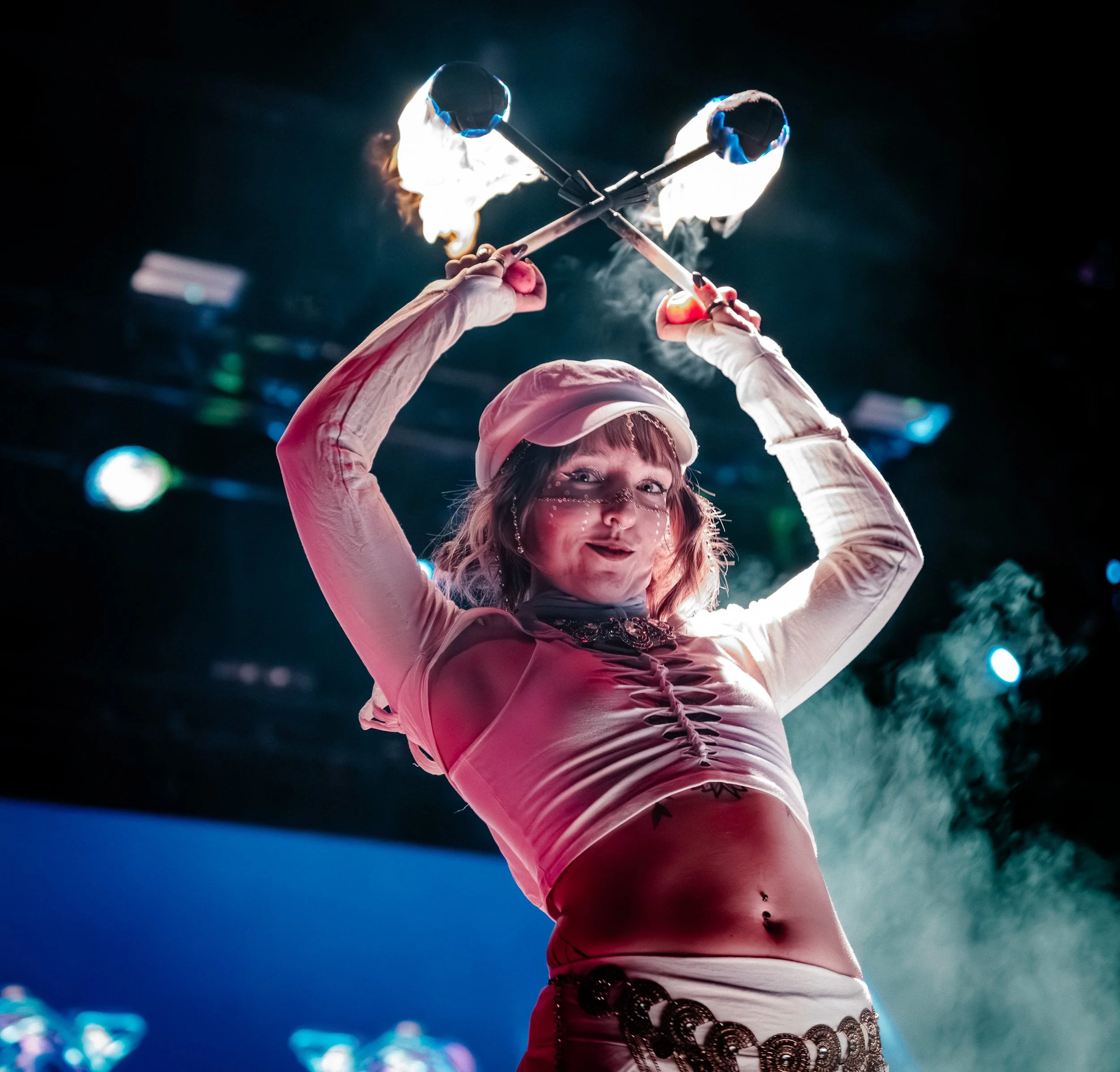 A female performer in a white outfit and hat twirling flaming sticks during a circus act.