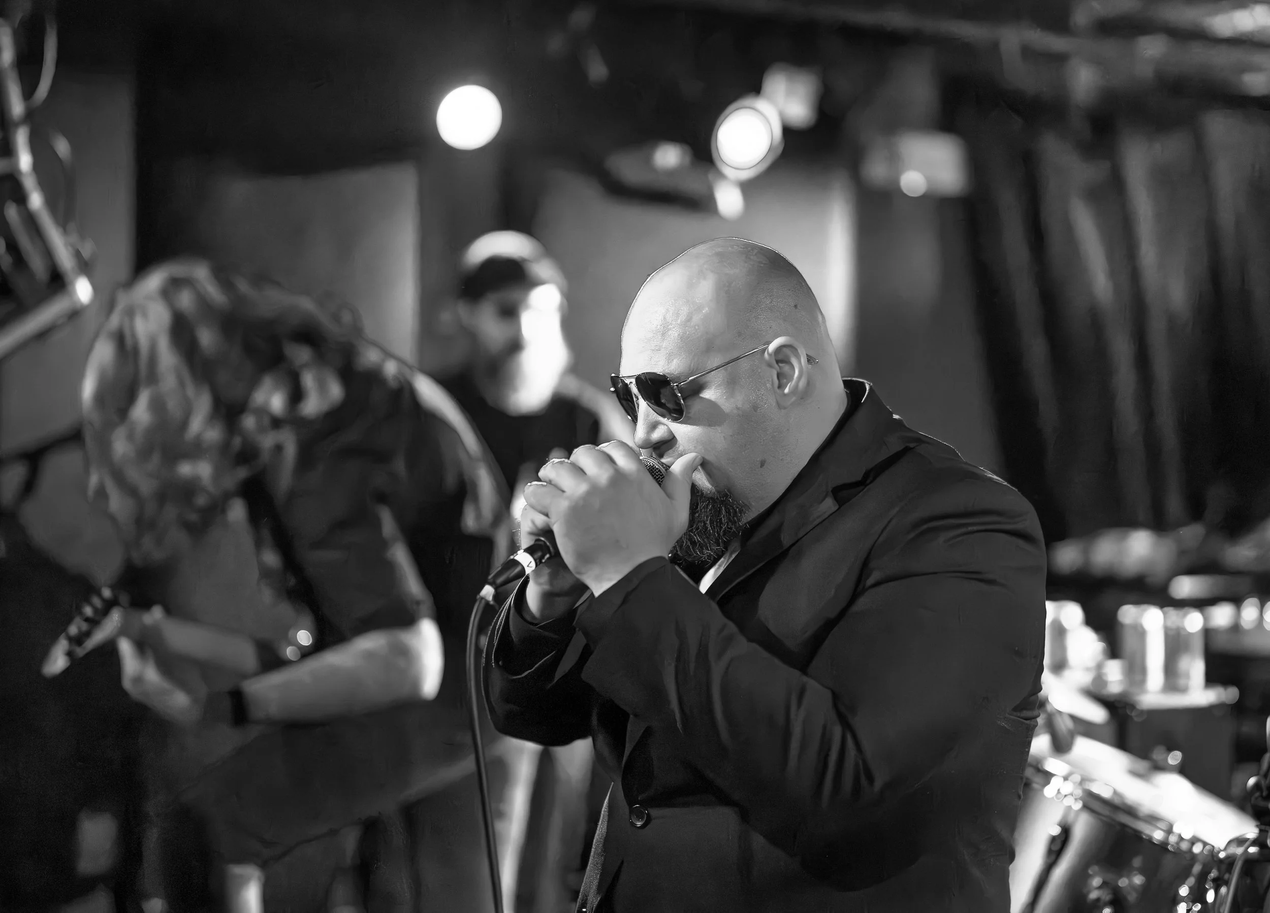 A bald man with sunglasses and a beard singing into a microphone, with a guitarist and a person wearing a face mask in the background, on a dimly lit stage.