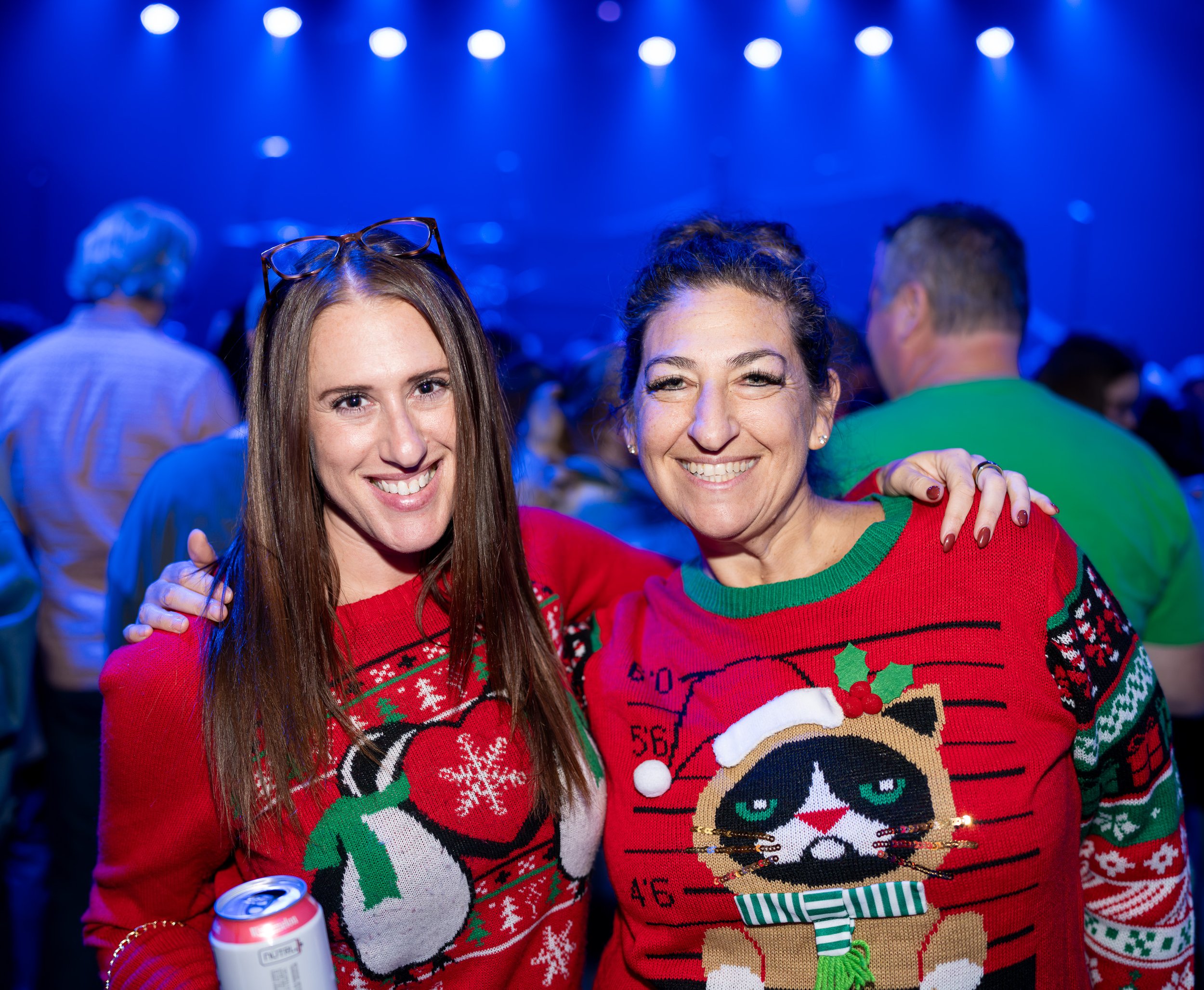 Two women smiling and wearing festive Christmas sweaters at a holiday party with a dark blue background and people in the crowd.