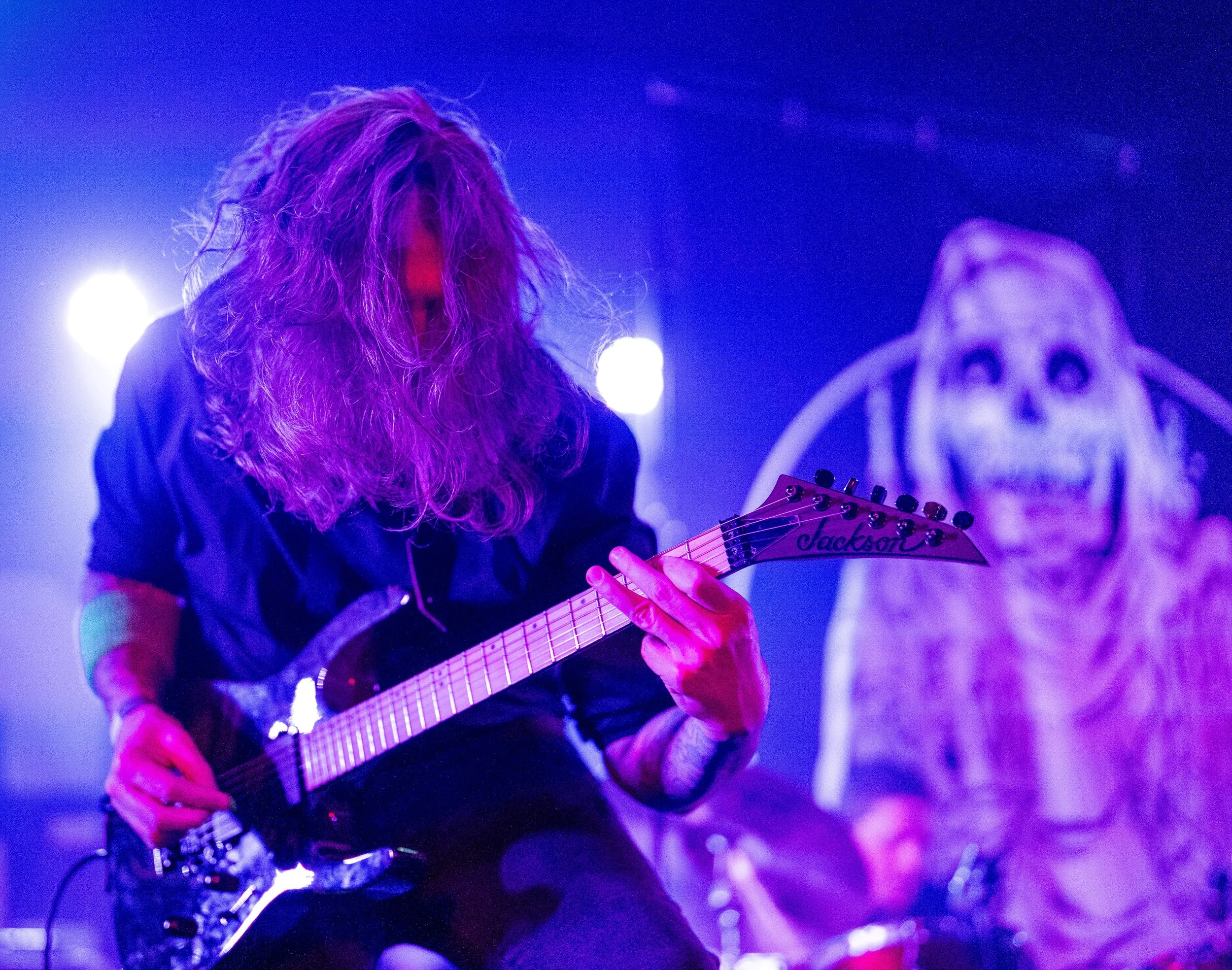 A long-haired guitarist performing on stage with purple and blue lighting, with a skeleton skull backdrop.
