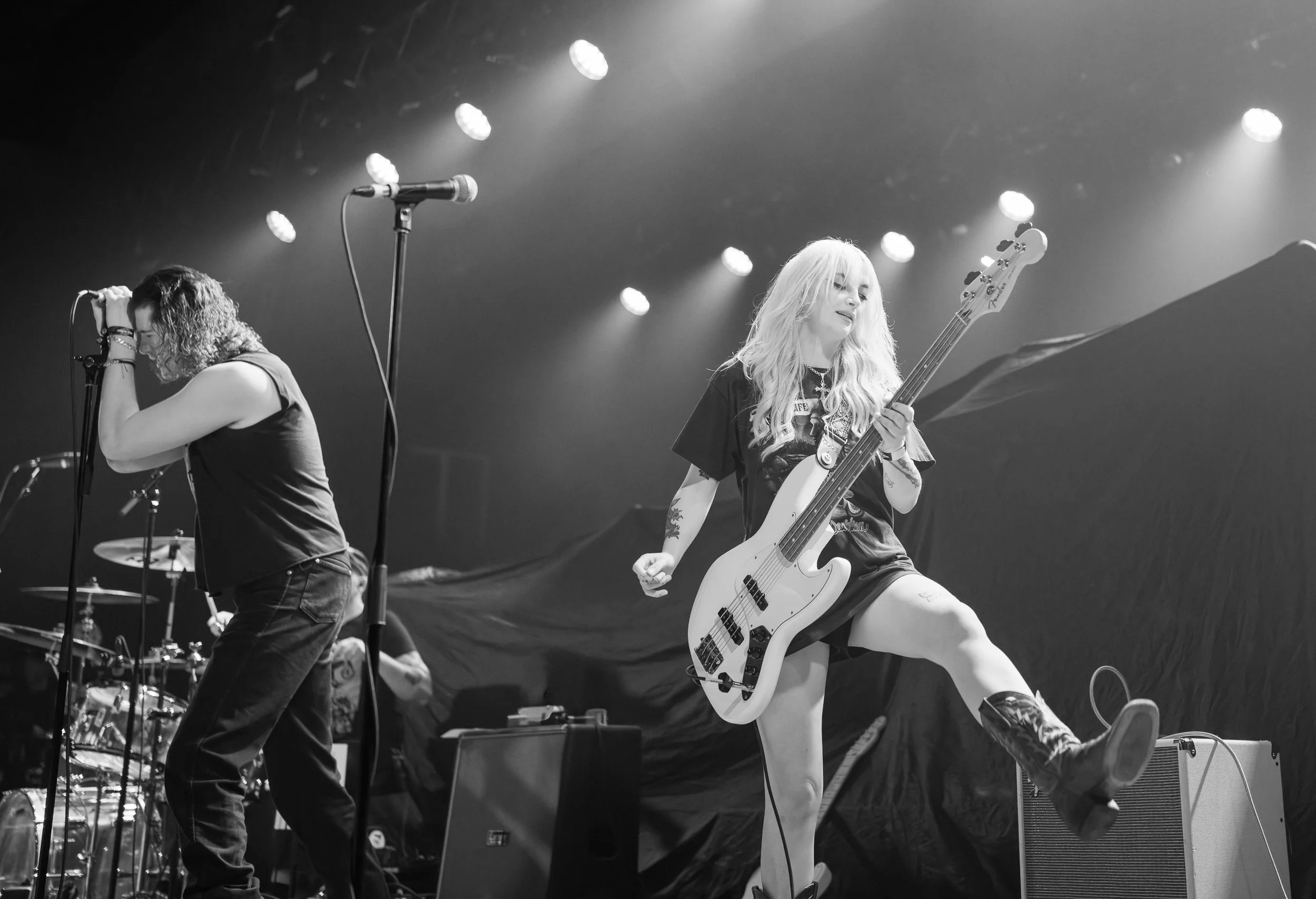 A black and white photo of a live music performance showing two female musicians on stage. One is singing into a microphone with curly hair, wearing a sleeveless top. The other is playing an electric bass guitar, with long blonde hair, wearing a t-sh