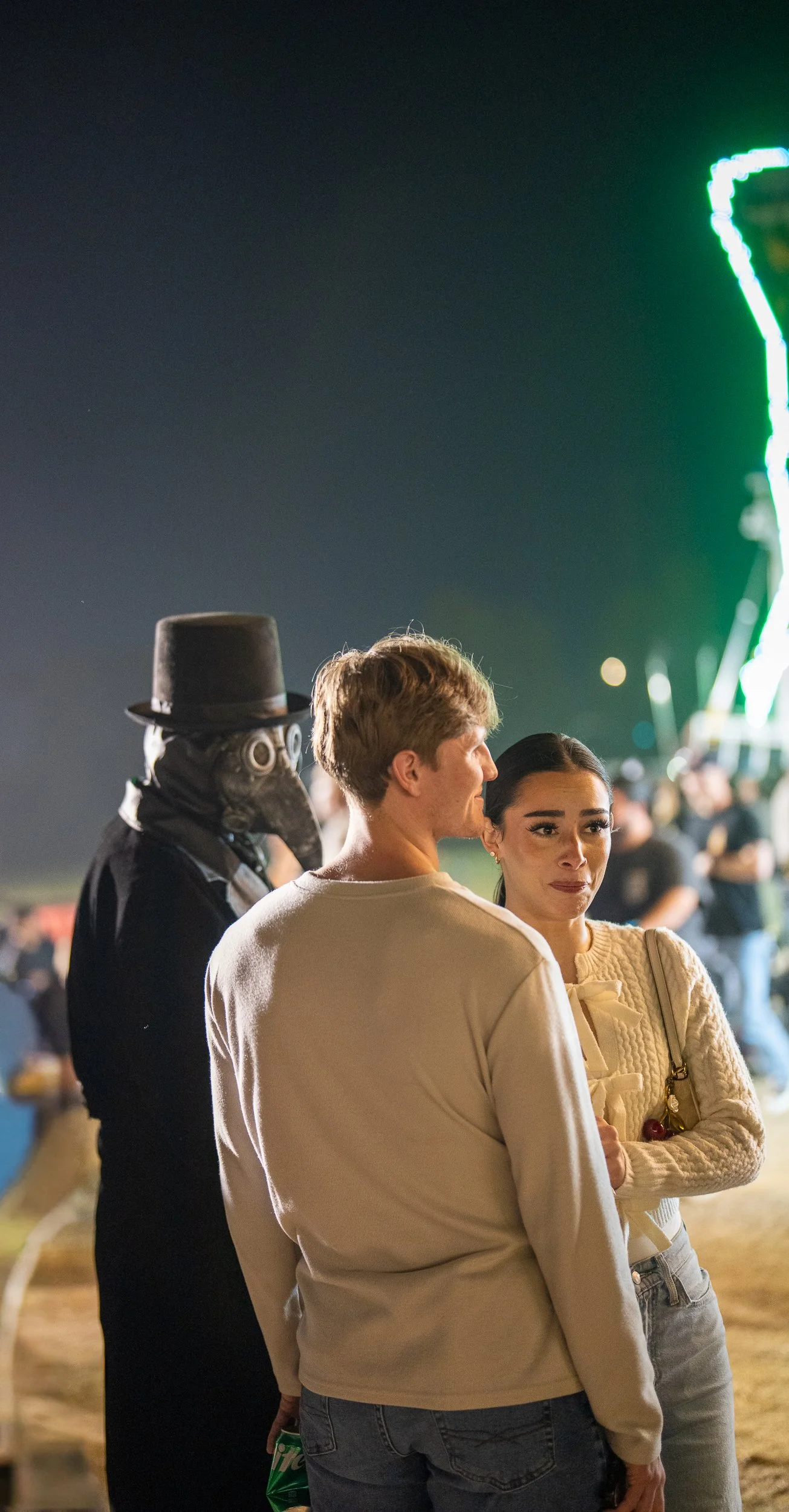 Two people standing close at night at a fairground, with a man wearing a plague doctor mask and a woman with a tearful expression, and brightly lit rides in the background.