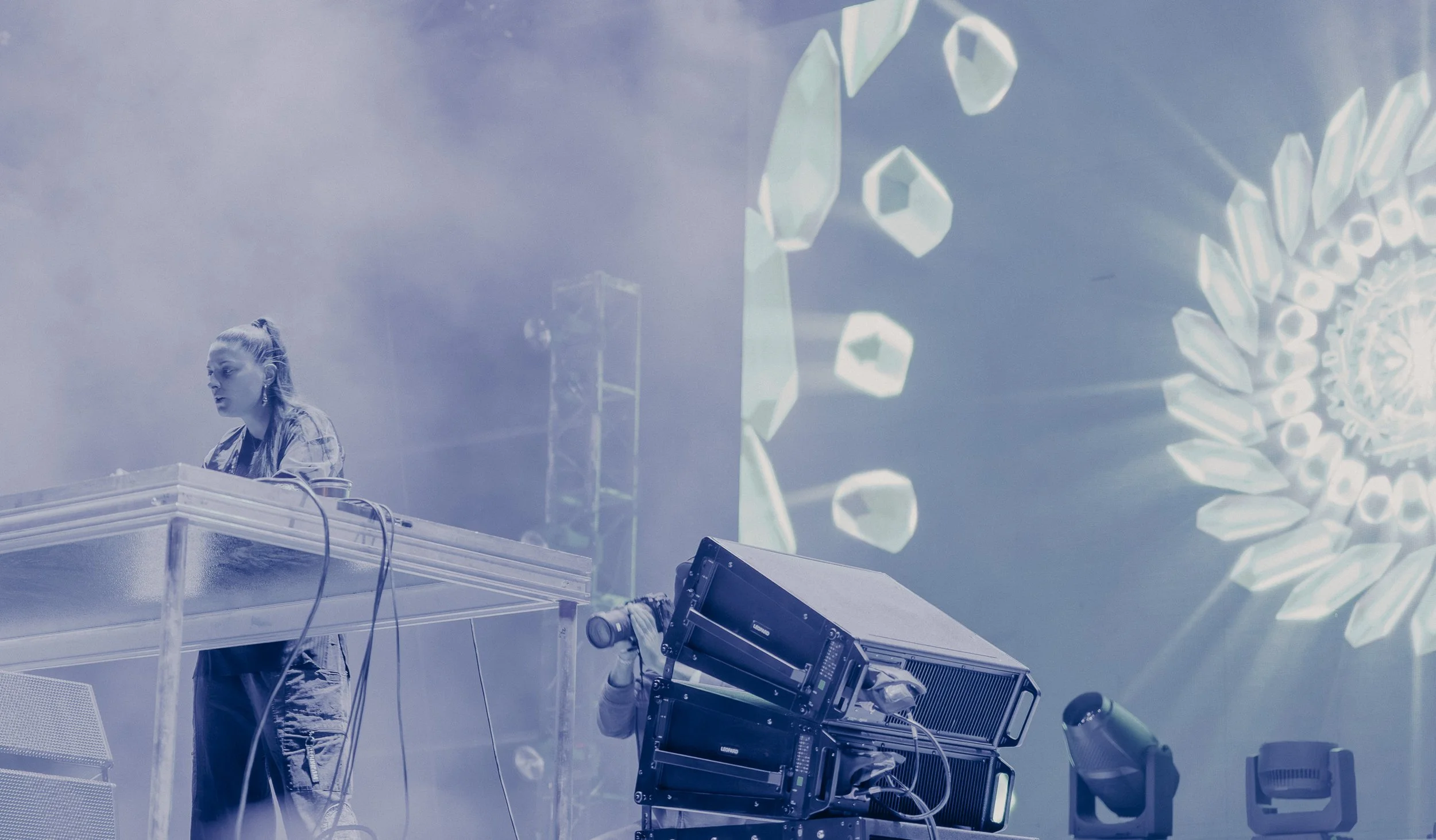 A female DJ performing on stage with electronic music equipment, with a large circular digital display in the background showing geometric shapes and patterns.
