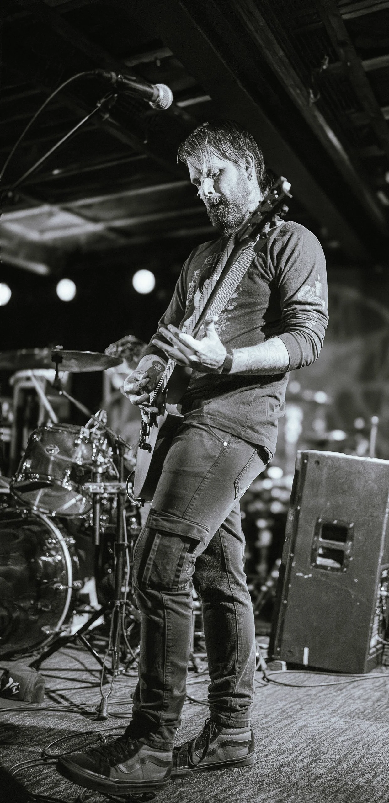 Black and white photo of a man playing an electric guitar on stage, with a drum set behind him, in a dimly lit venue.