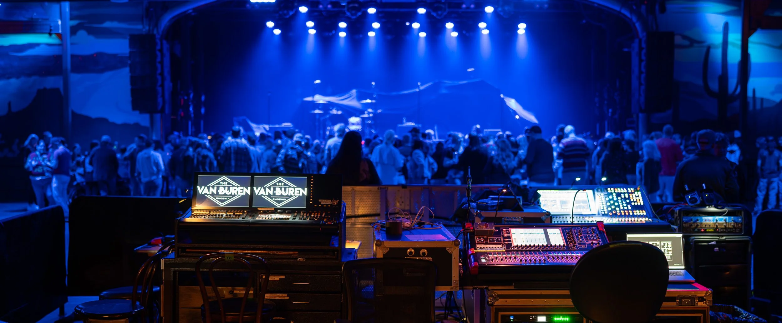A stage with vibrant blue lighting and a crowd of people gathering, seen from behind a DJ booth with music equipment and monitors, at a concert venue.