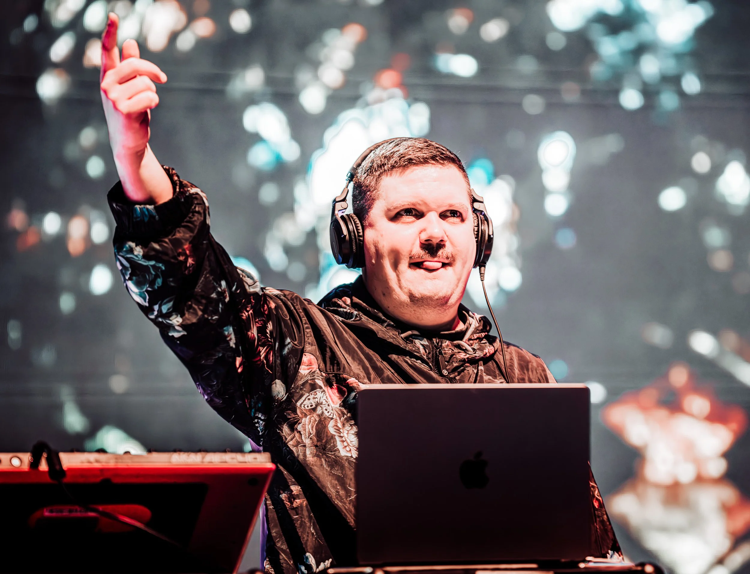 A DJ wearing headphones and a patterned jacket, performing at a concert or event with a blurred light background.