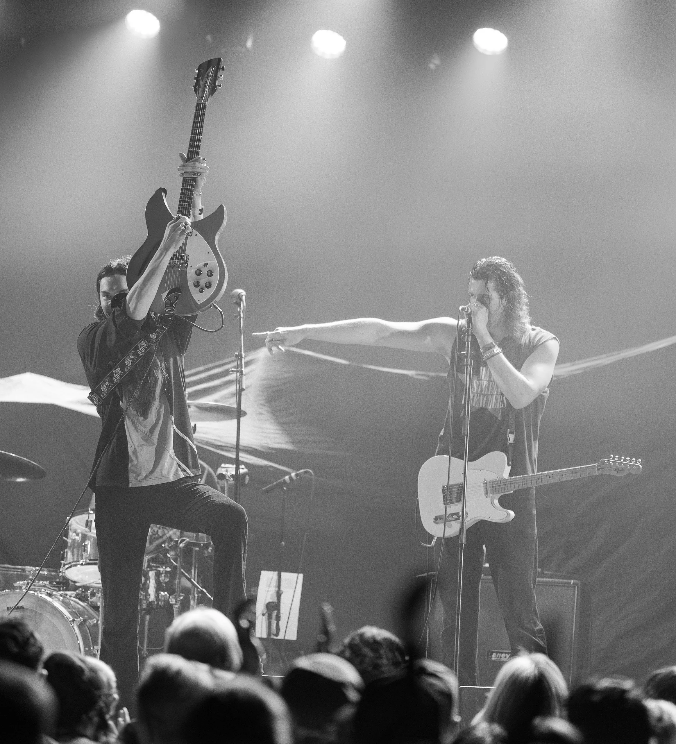 Black and white photo of a live music concert with two female musicians on stage; one is holding a guitar in the air, and the other is singing into a microphone while pointing at her, with an electric guitar hanging from her shoulder; audience member