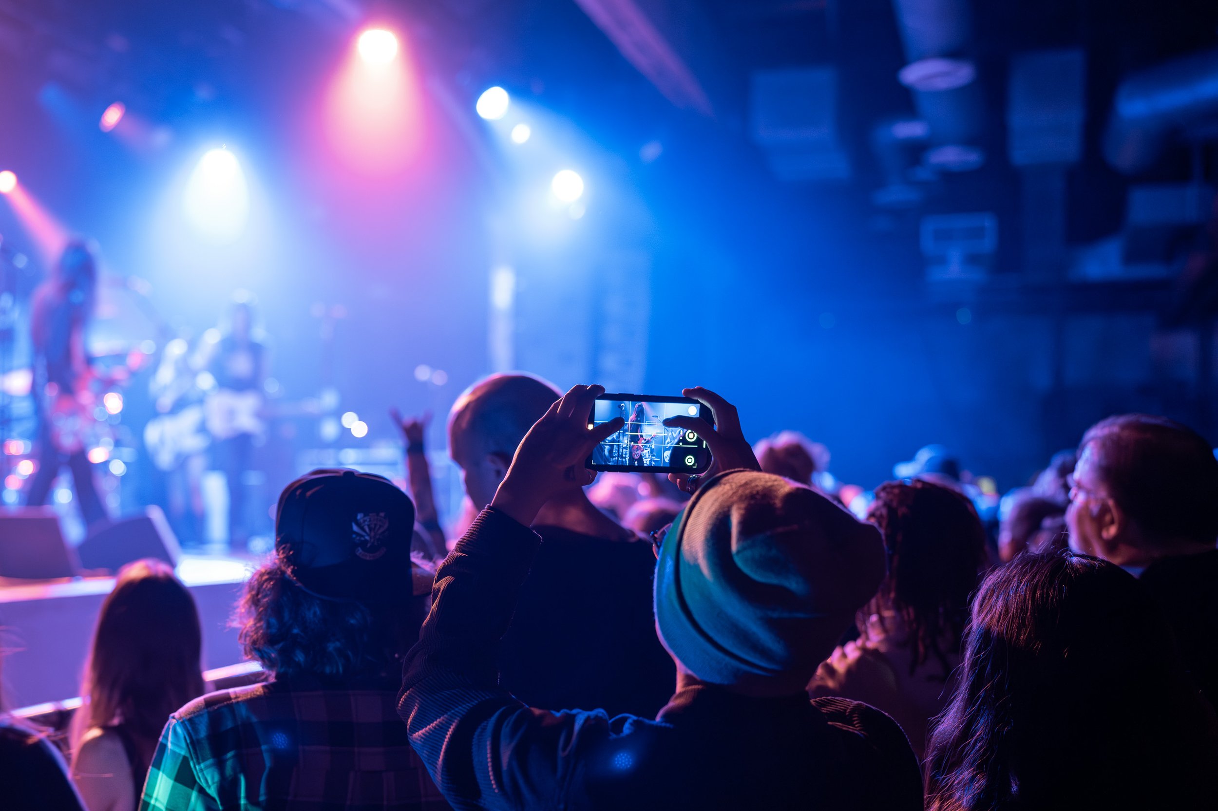 Audience watching a concert with colorful stage lighting, one person capturing the performance on a smartphone.