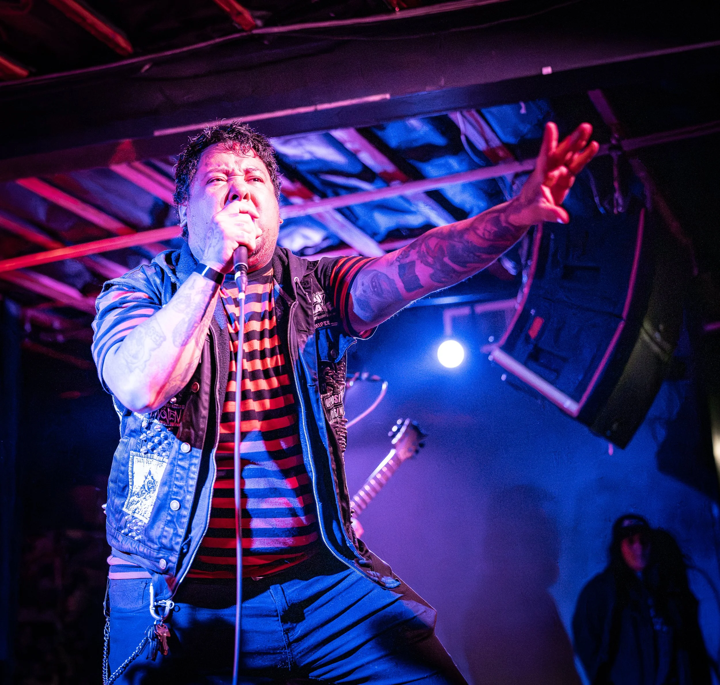 Male singer performing on stage with microphone, wearing a striped shirt and vest, with tattoos on arm, under stage lighting.