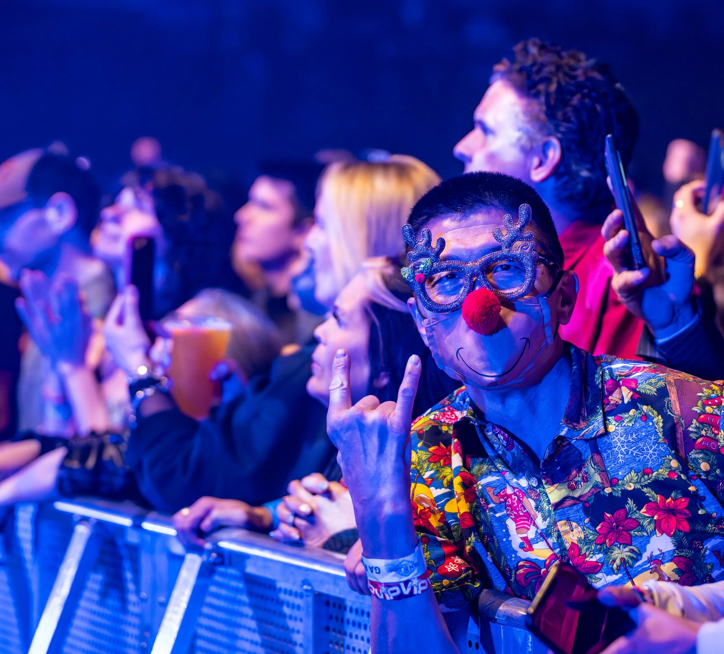 A crowd of people at a concert or event, with one man in the foreground wearing festive glasses with reindeer antlers, a red nose, a colorful Hawaiian shirt, and making the rock on hand gesture.