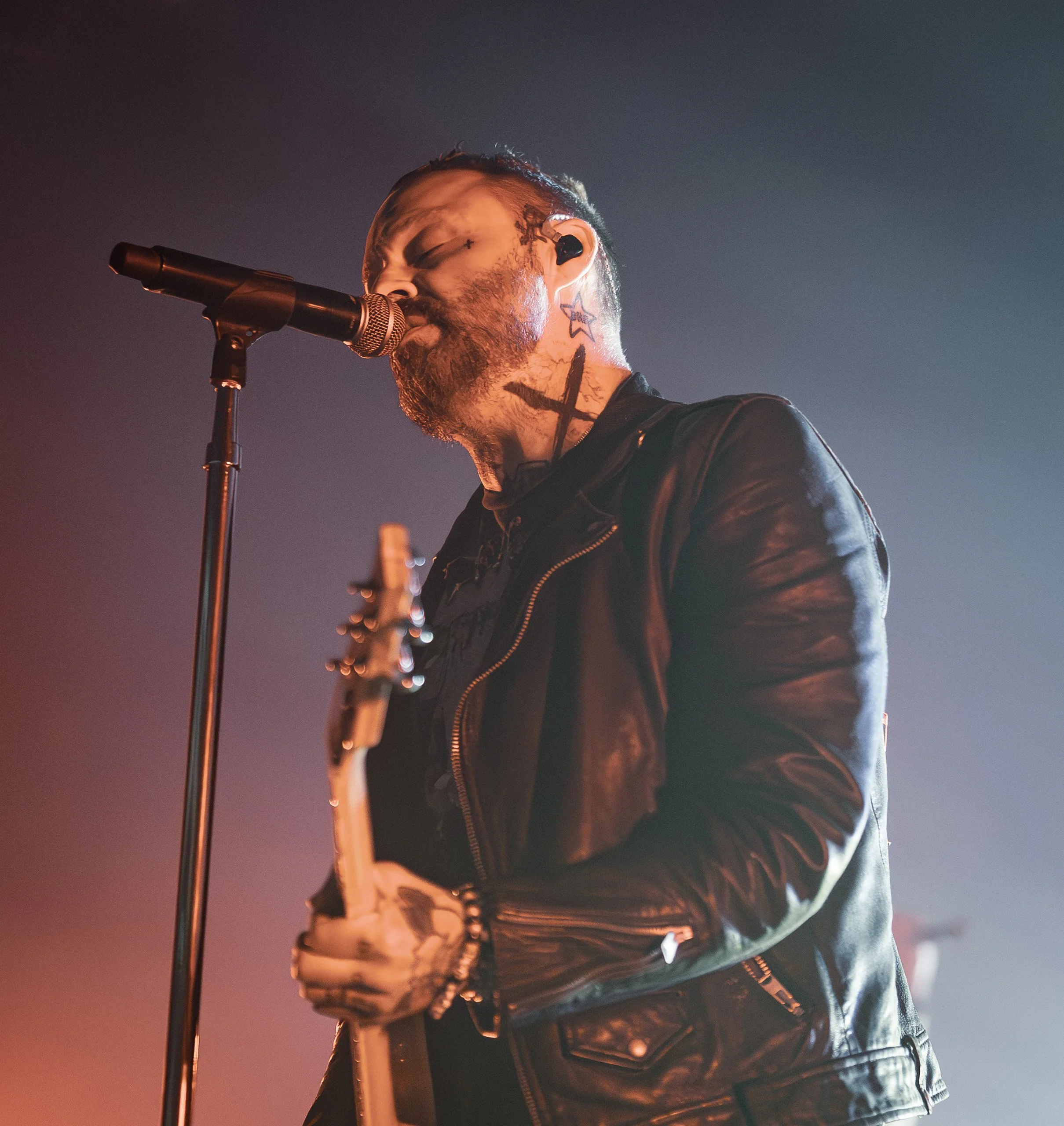 A male musician with facial tattoos singing into a microphone while playing an electric guitar, wearing a black leather jacket, on stage with colorful lighting