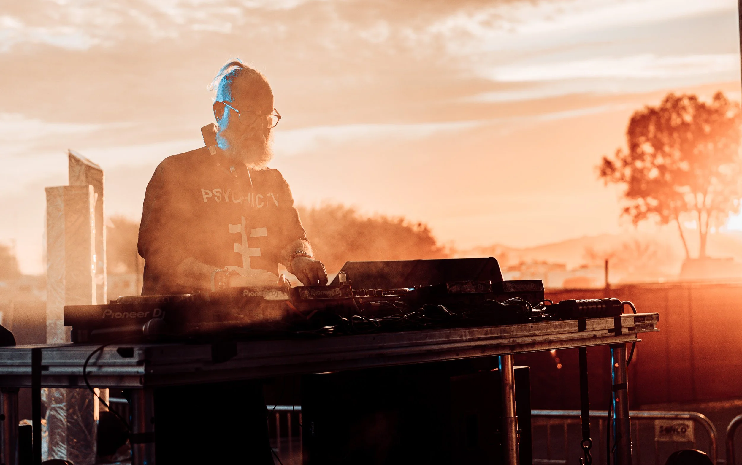 A DJ with a gray beard and glasses performing outdoors during a sunset, wearing a black hoodie with the word the word 'PSYCHIC' on it, surrounded by warm orange lighting.