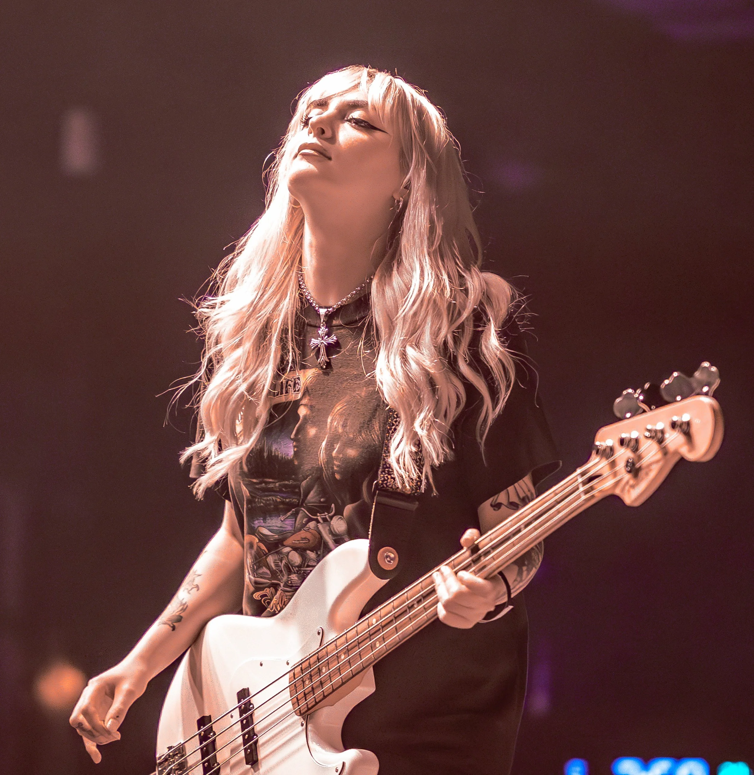 A woman with long blonde wavy hair playing a white electric bass guitar on stage, wearing a graphic t-shirt and jewelry.