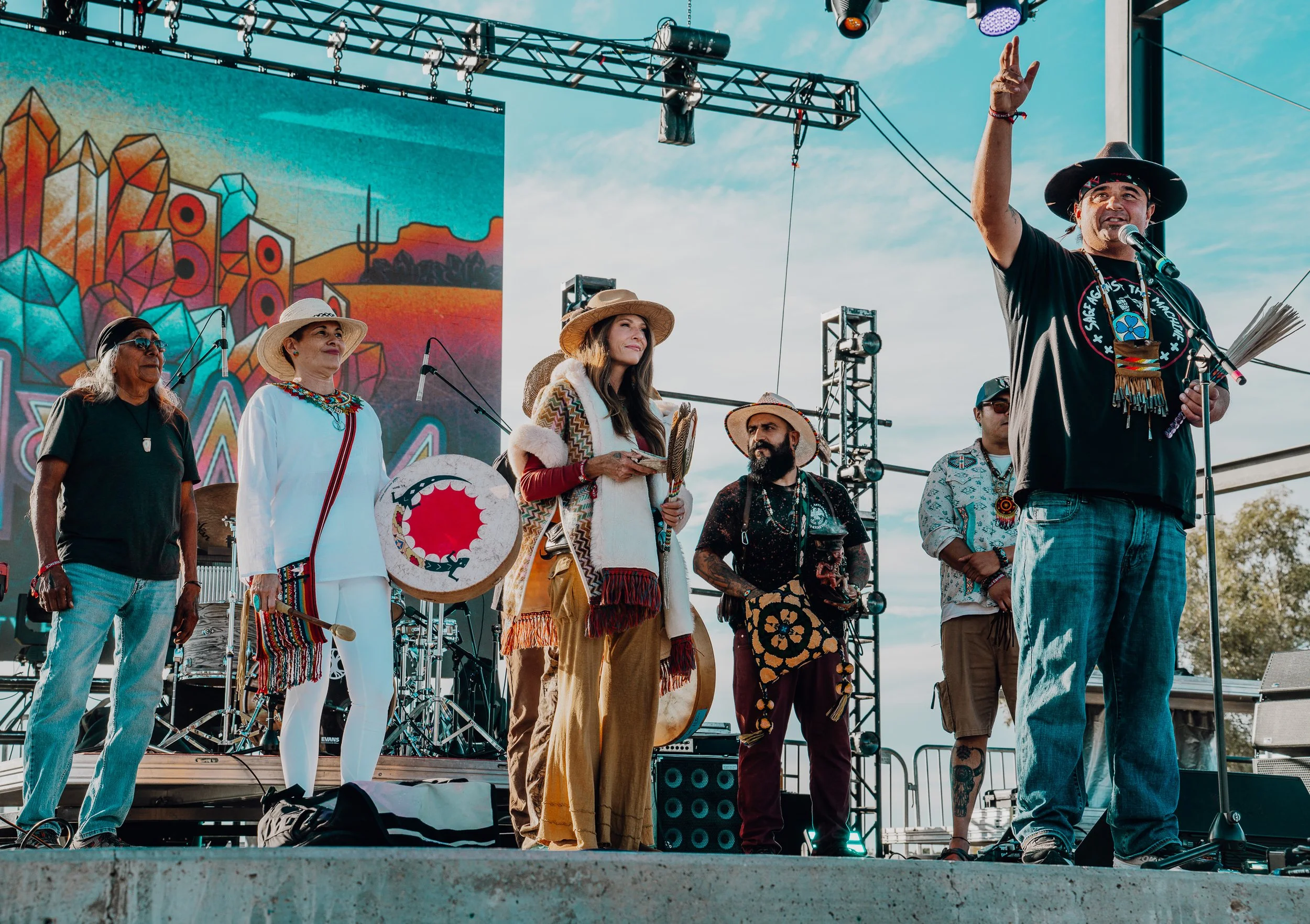 A group of diverse people on stage at an outdoor cultural event, with some holding traditional items, a man speaking at a microphone, and a colorful mural in the background.
