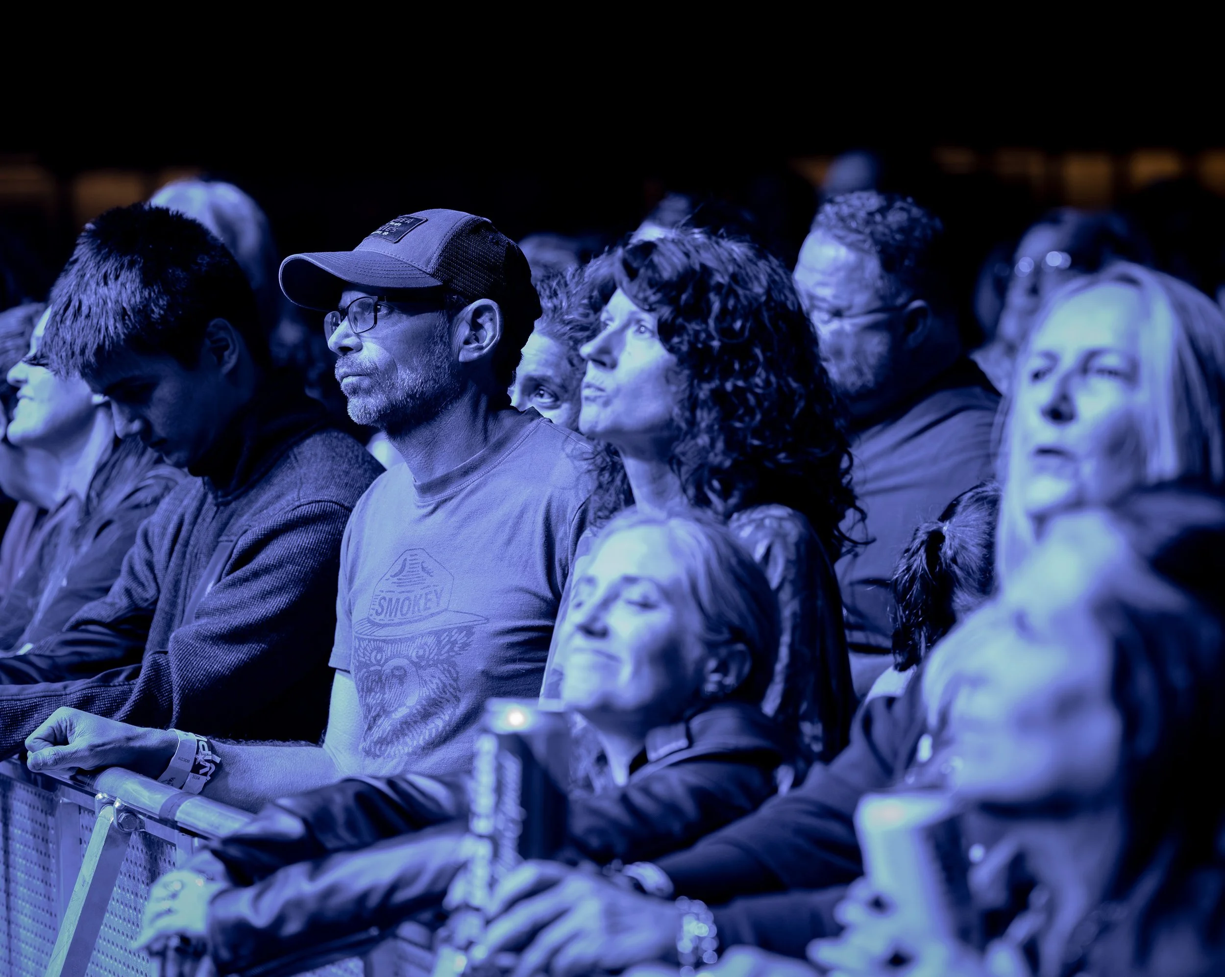 Audience members sitting at a concert or event, illuminated by blue lighting, paying attention to the stage.