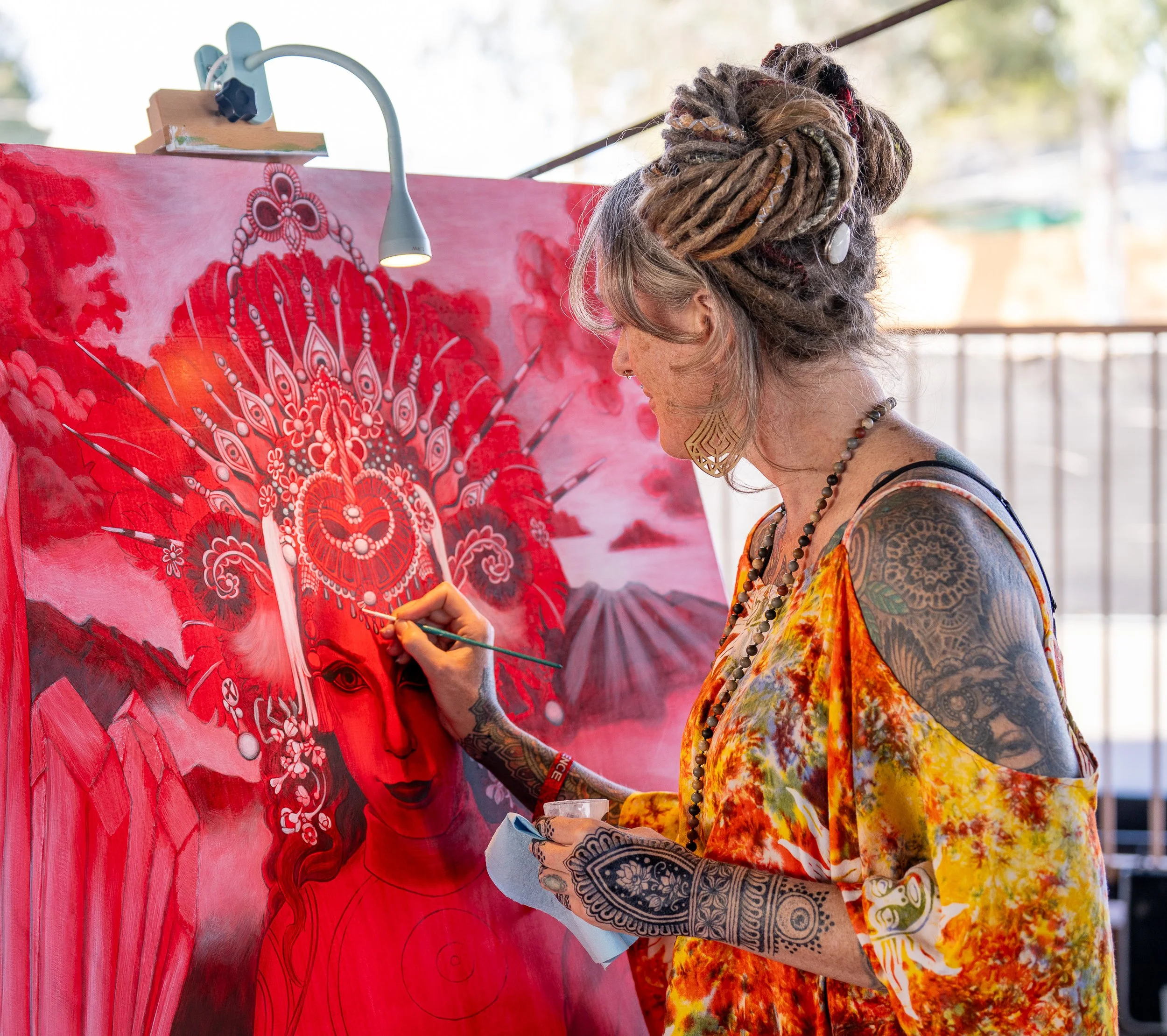 A woman with tattoos and dreadlocks painting on a large canvas. She is dressed in a colorful, patterned dress and is focused on her artwork, which features a stylized portrait of a woman with intricate headdress in shades of red and pink.