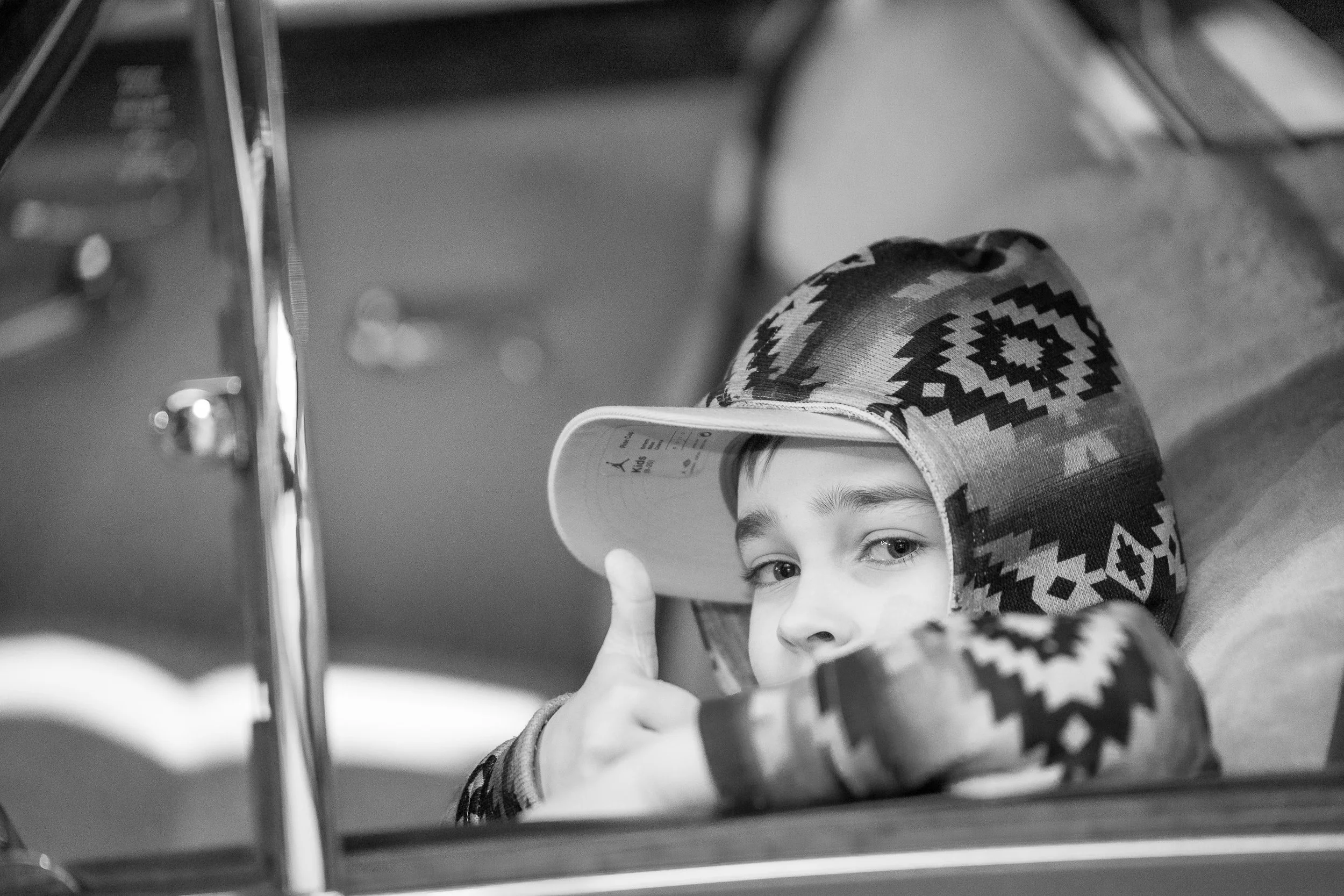 A young boy wearing a patterned hoodie and a baseball cap giving a thumbs-up while leaning on a surface, with a reflective metal object in the foreground.
