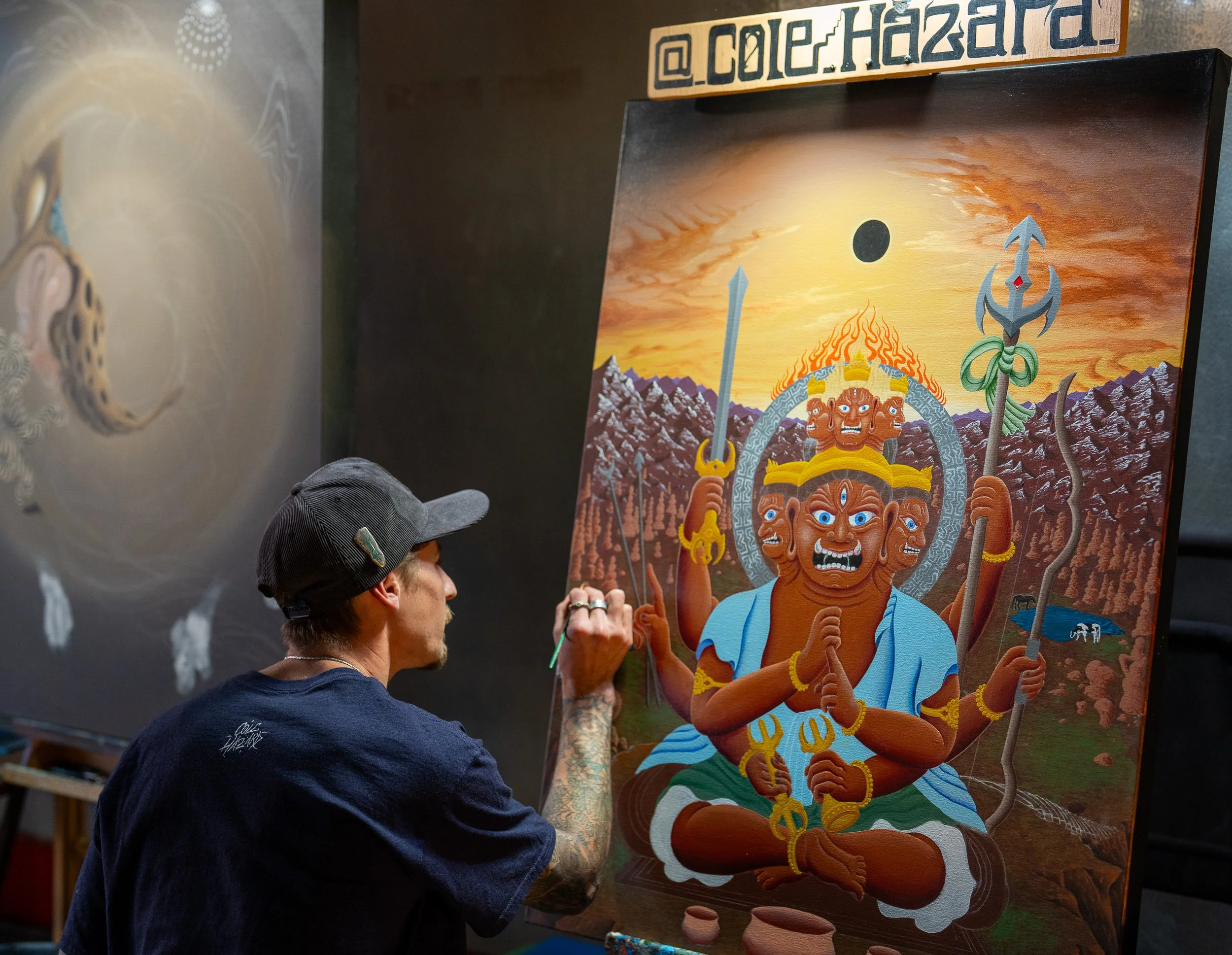 An artist painting a colorful depiction of a multi-armed deity with multiple faces, holding various objects, against a mountain landscape and a sunset sky. The artist is wearing a dark t-shirt and a black baseball cap, focused on the painting.