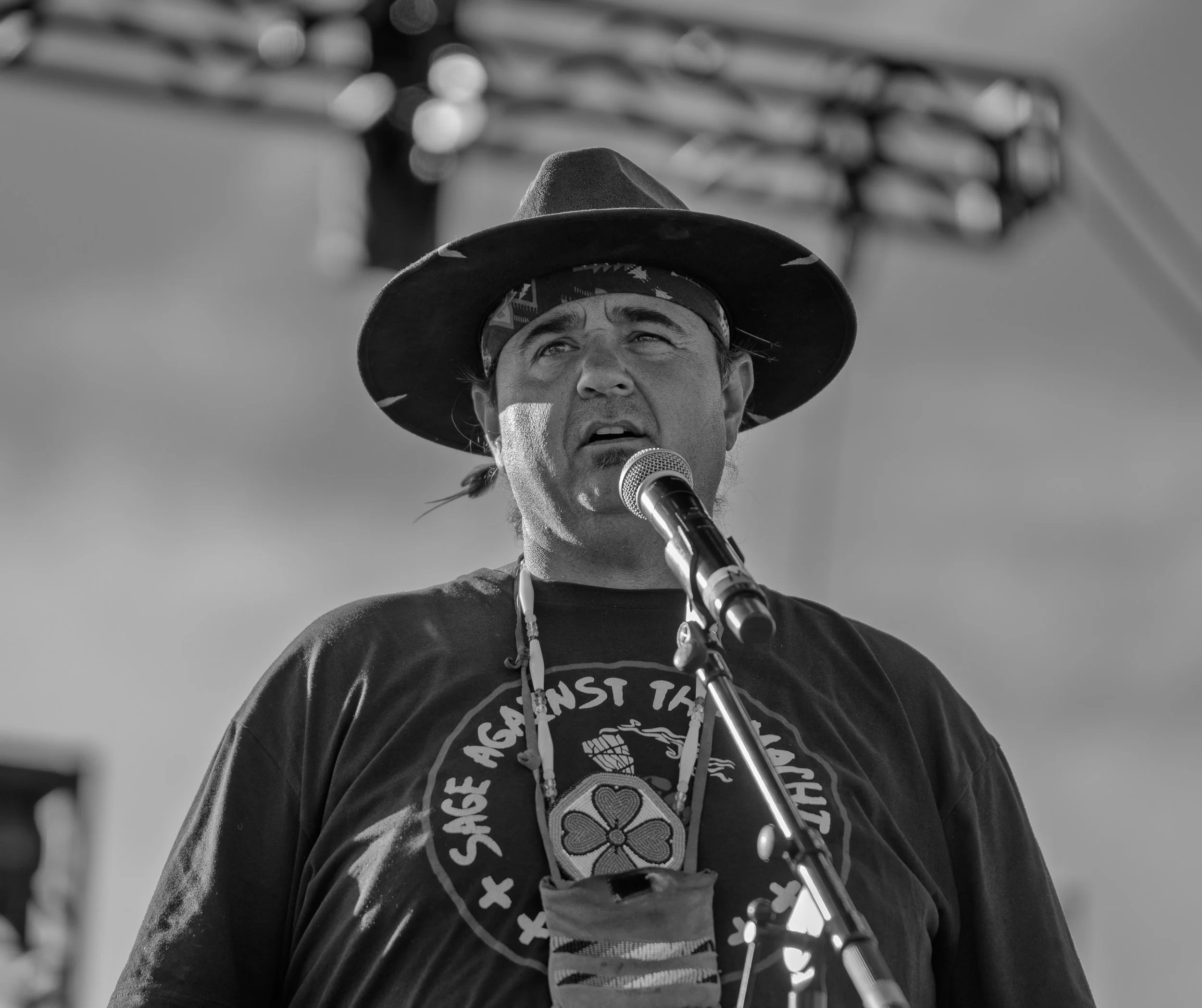 A man wearing a wide-brimmed hat and a bandana sings into a microphone during a performance.