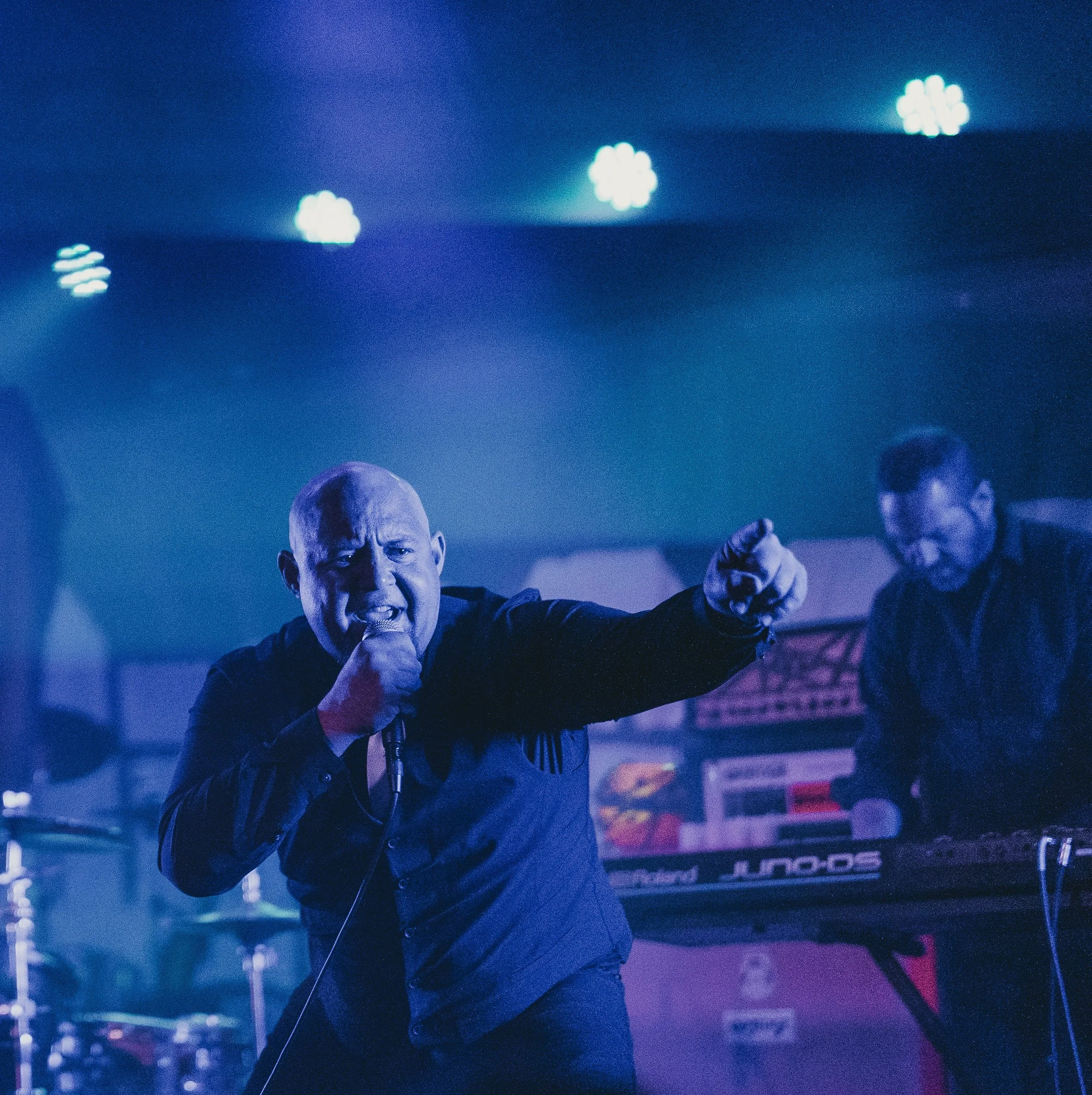 A man passionately singing or shouting into a microphone on stage with a keyboard player in the background, under blue stage lighting.