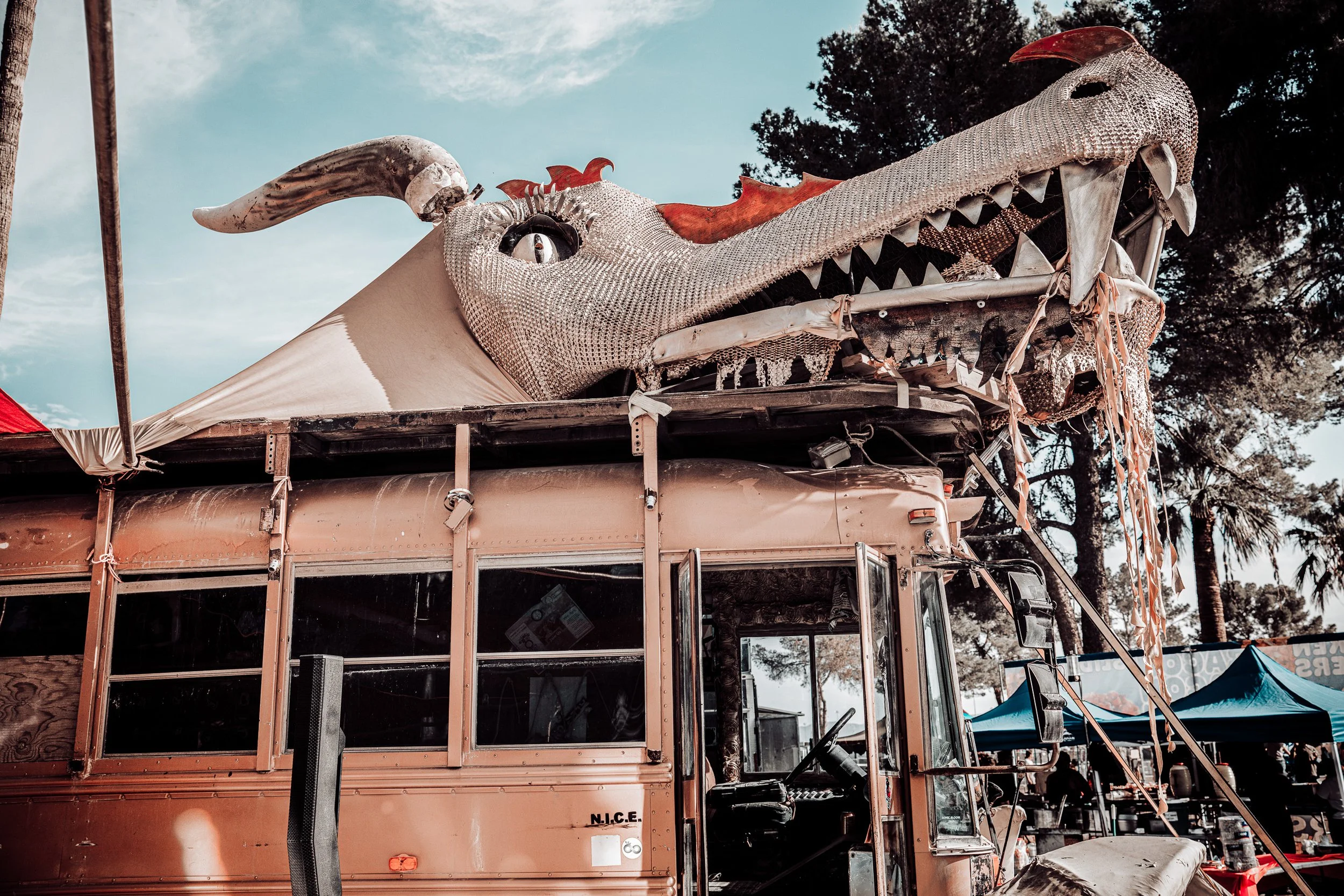 A large dragon head sculpture mounted on top of a bus, with the dragon's mouth open. The sculpture appears to be made of metallic or reflective materials and has red accents on the crest and horns. The bus is parked outdoors with tents and trees visi