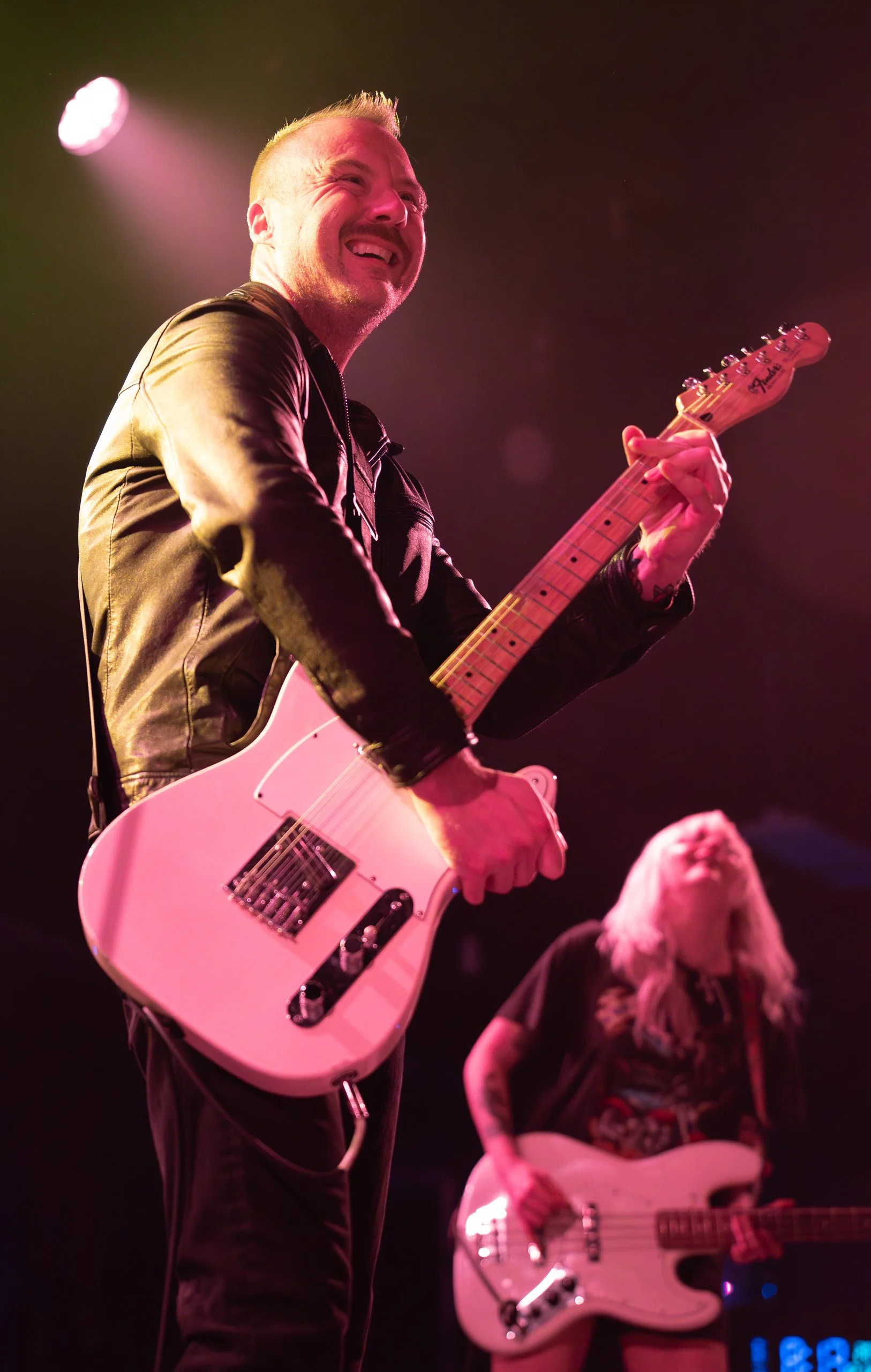Musician playing a white electric guitar on stage, smiling with a leather jacket, pink stage lighting.