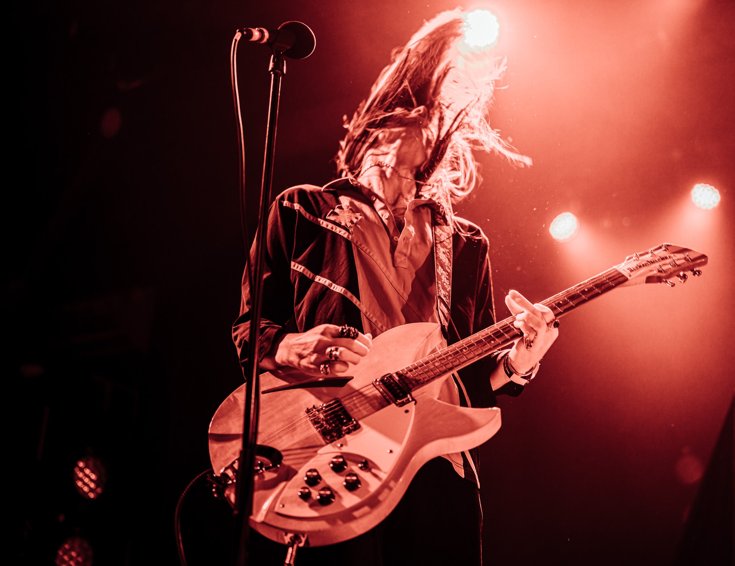 A female musician plays an electric guitar on stage, with long hair and wearing a jacket, under warm red stage lighting.