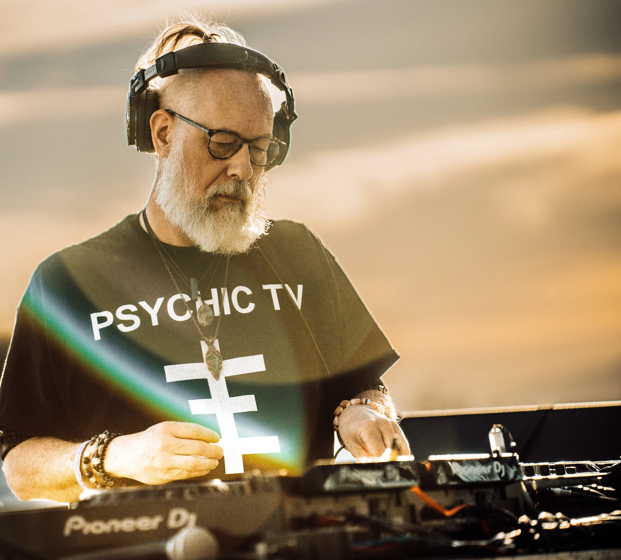 Older man with white beard and glasses wearing headphones, focused on DJ equipment, wearing a black T-shirt that reads 'PSYCHIC TV' with a large cross symbol.