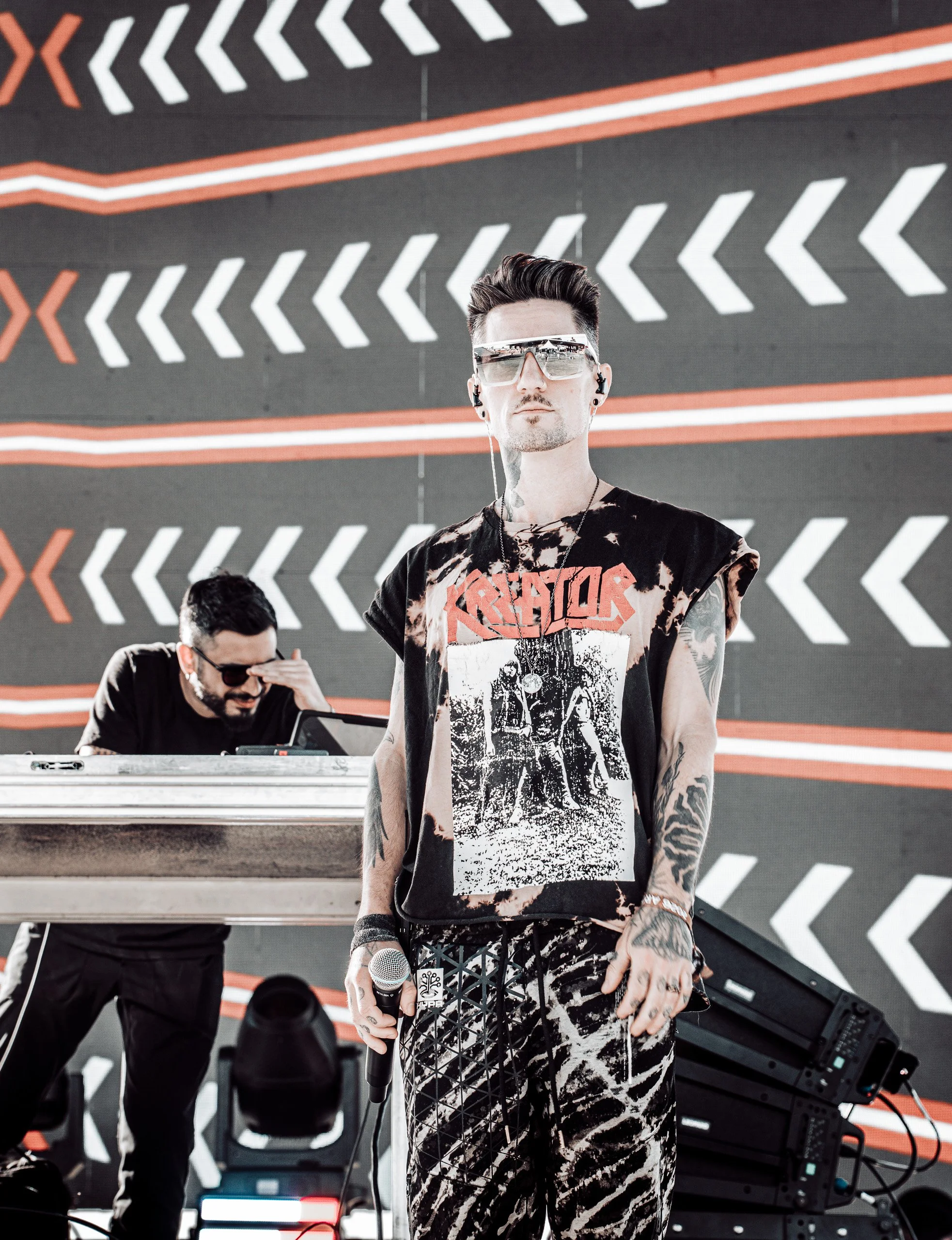 A man wearing sunglasses, a torn graphic T-shirt, and patterned pants holding a microphone stands in front of a DJ at a music event, with a background of geometric patterns on a wall.
