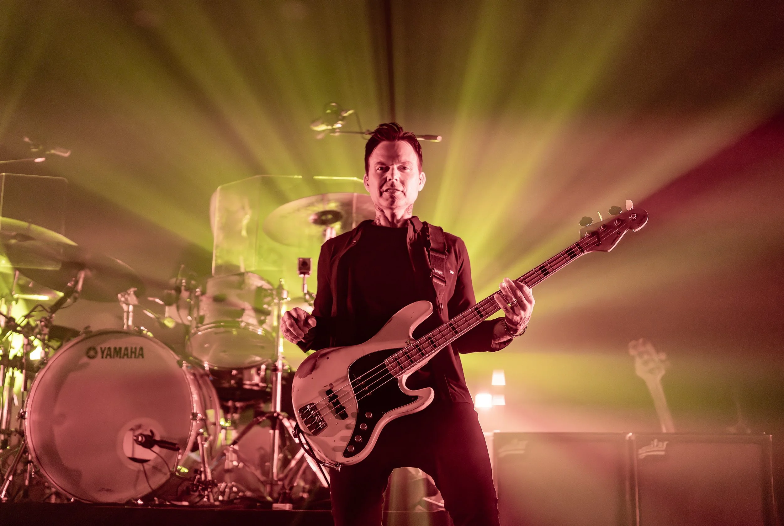 A man playing a bass guitar on stage, with a drummer and drum set in the background, illuminated by yellow stage lighting.