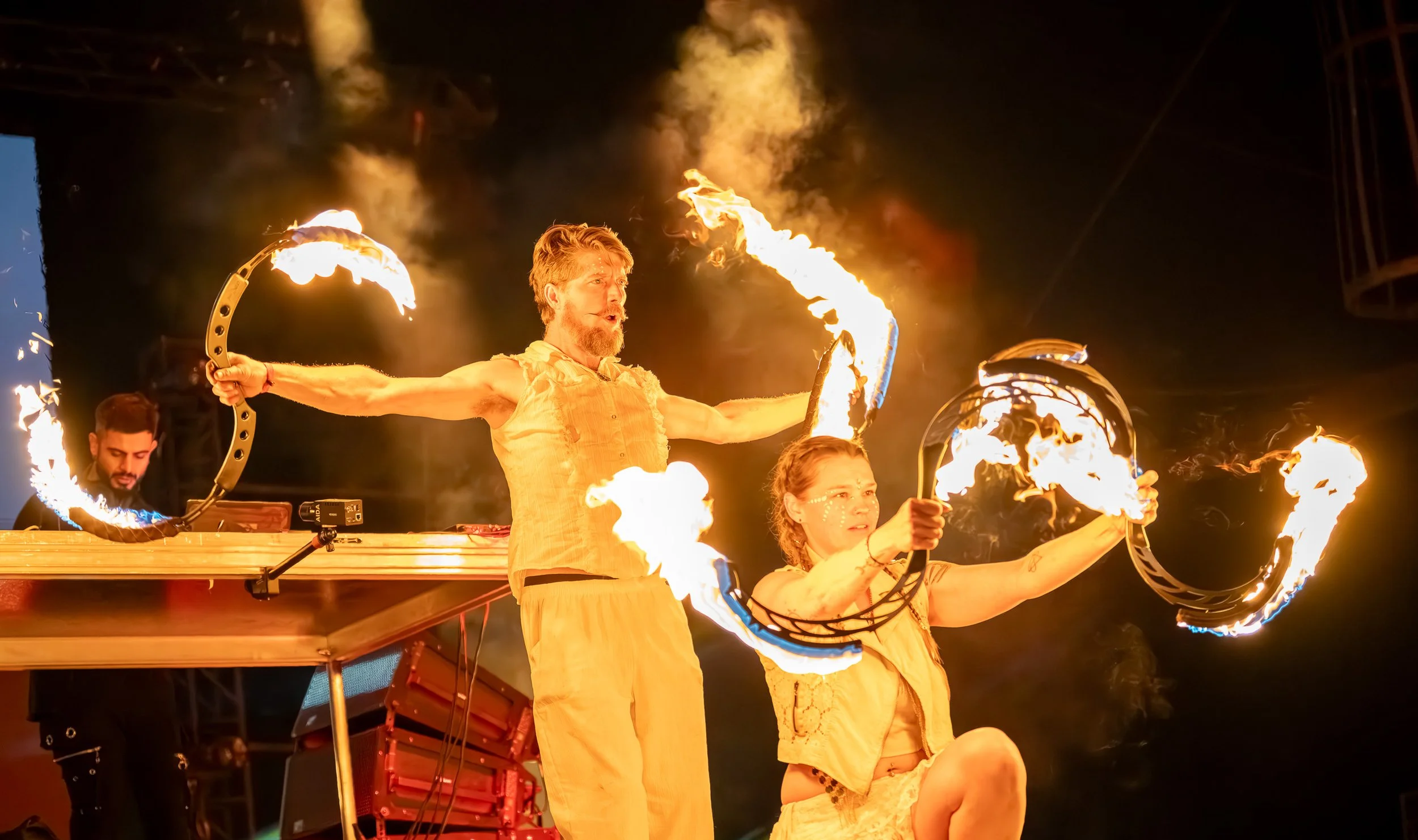 Two performers holding fire torches during a nighttime fire show on stage, with a DJ in the background.