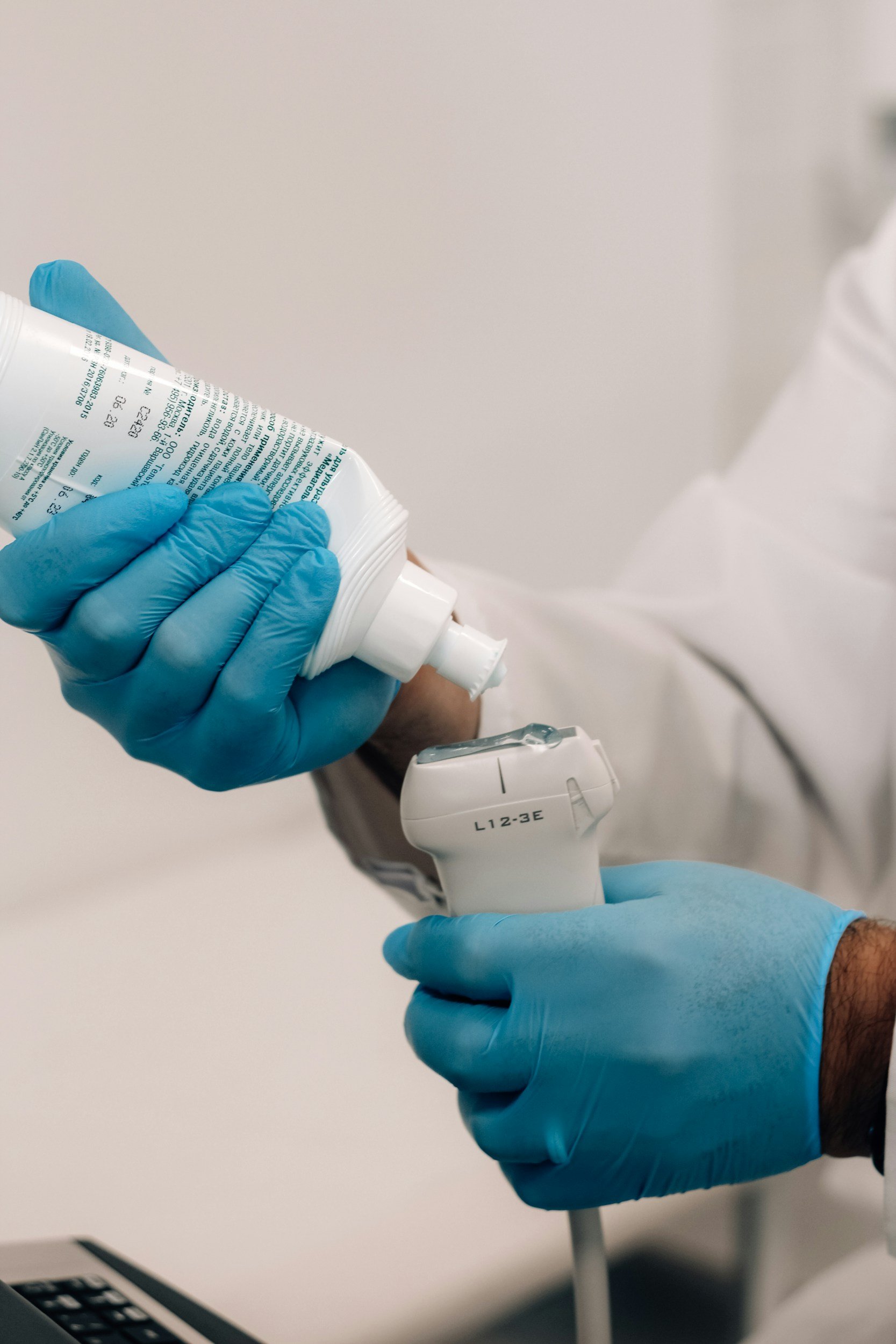 A person wearing blue gloves holding a barcode scanner and a bottle of liquid, possibly hand sanitizer or disinfectant, in a clinical or medical setting.