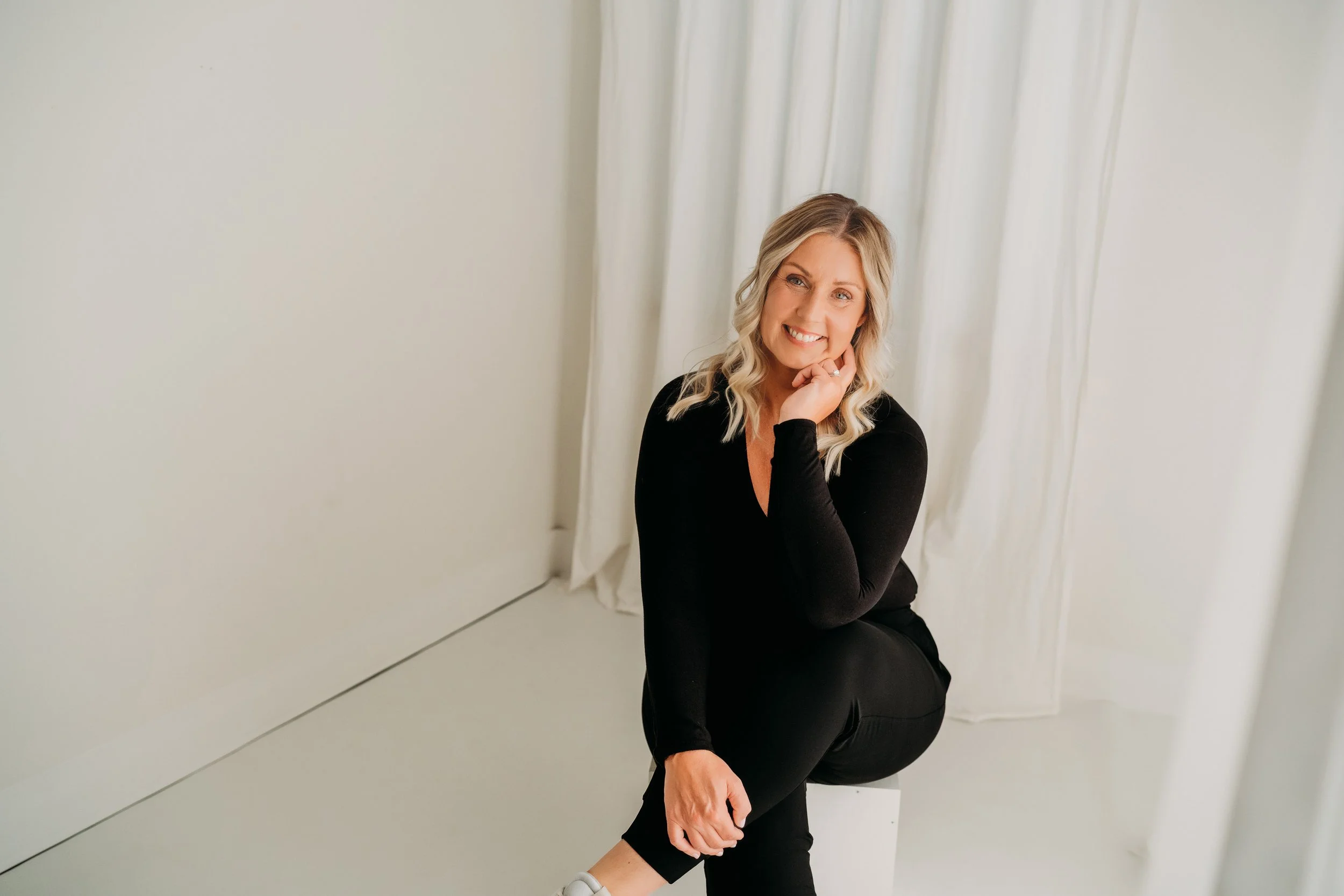 A woman sitting on a white cube, wearing black clothing, smiling, with long blonde hair, in front of white curtains and wall.