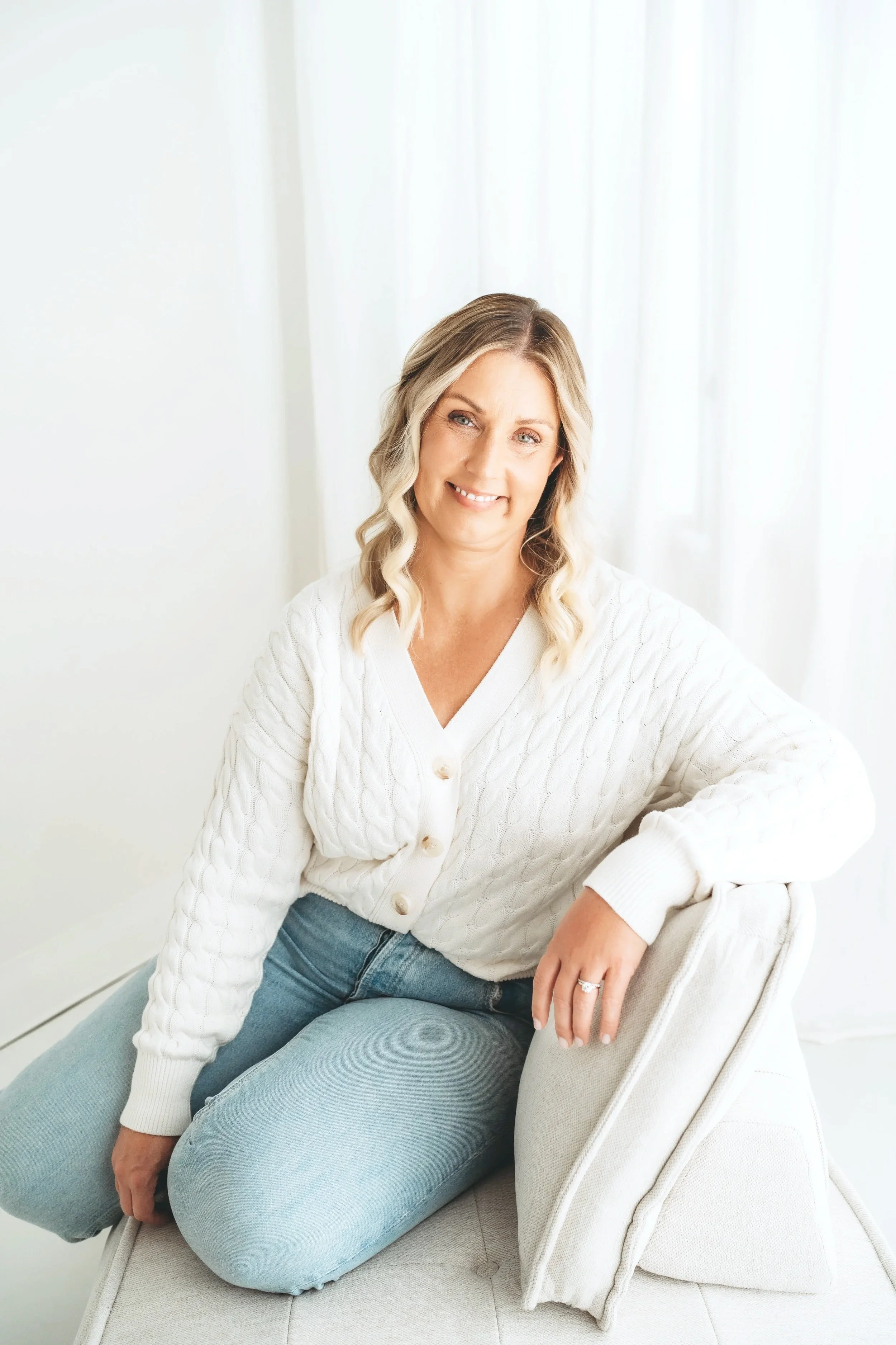 A woman with blonde wavy hair wearing a white cable knit cardigan and light blue jeans, sitting on a beige sofa in a bright room with white curtains.