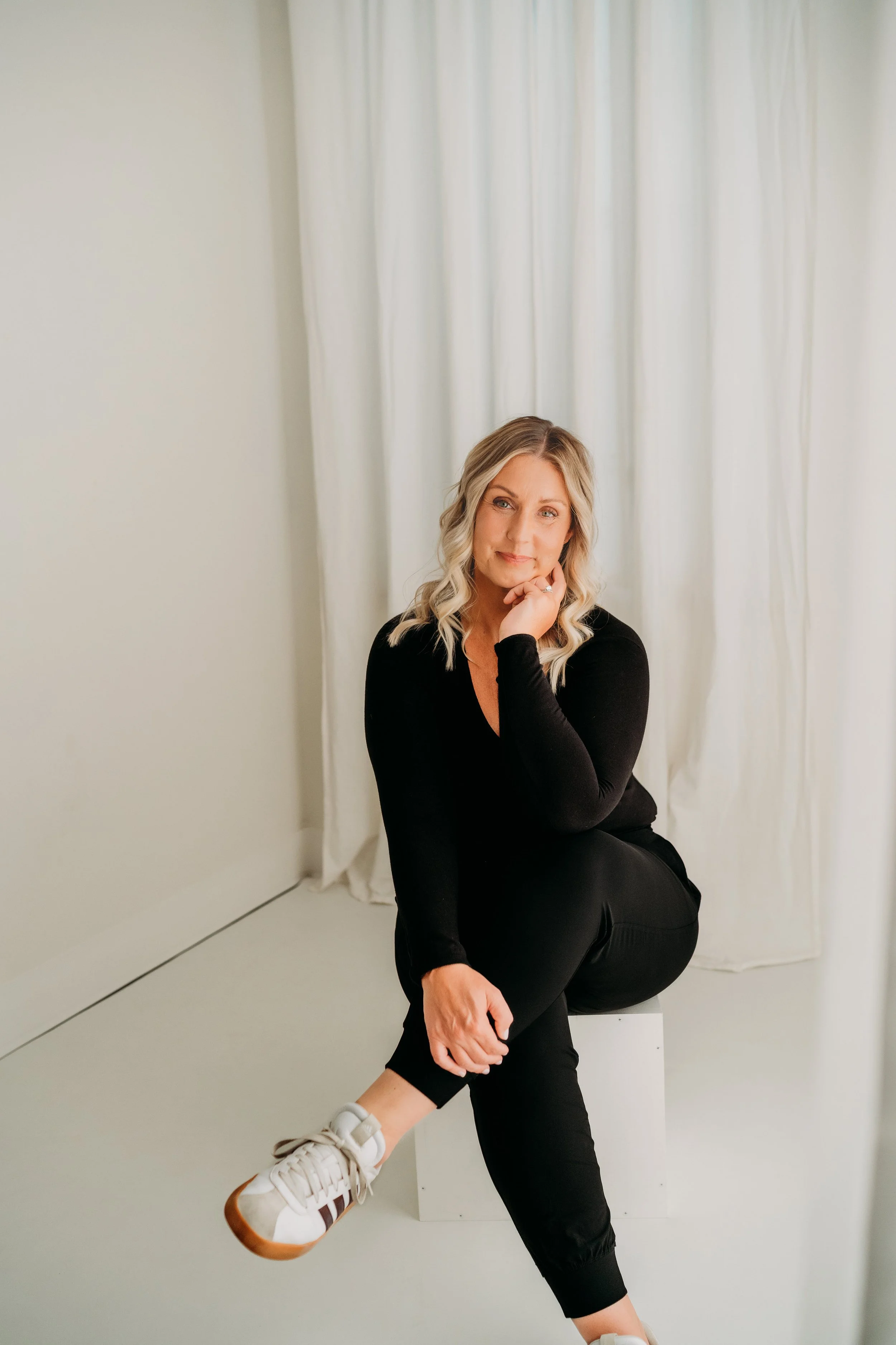 A woman with blonde hair in loose waves, wearing a black long-sleeven top and black pants, sitting on a block in front of white curtains, looking at the camera with a slight smile, resting her chin on her hand.