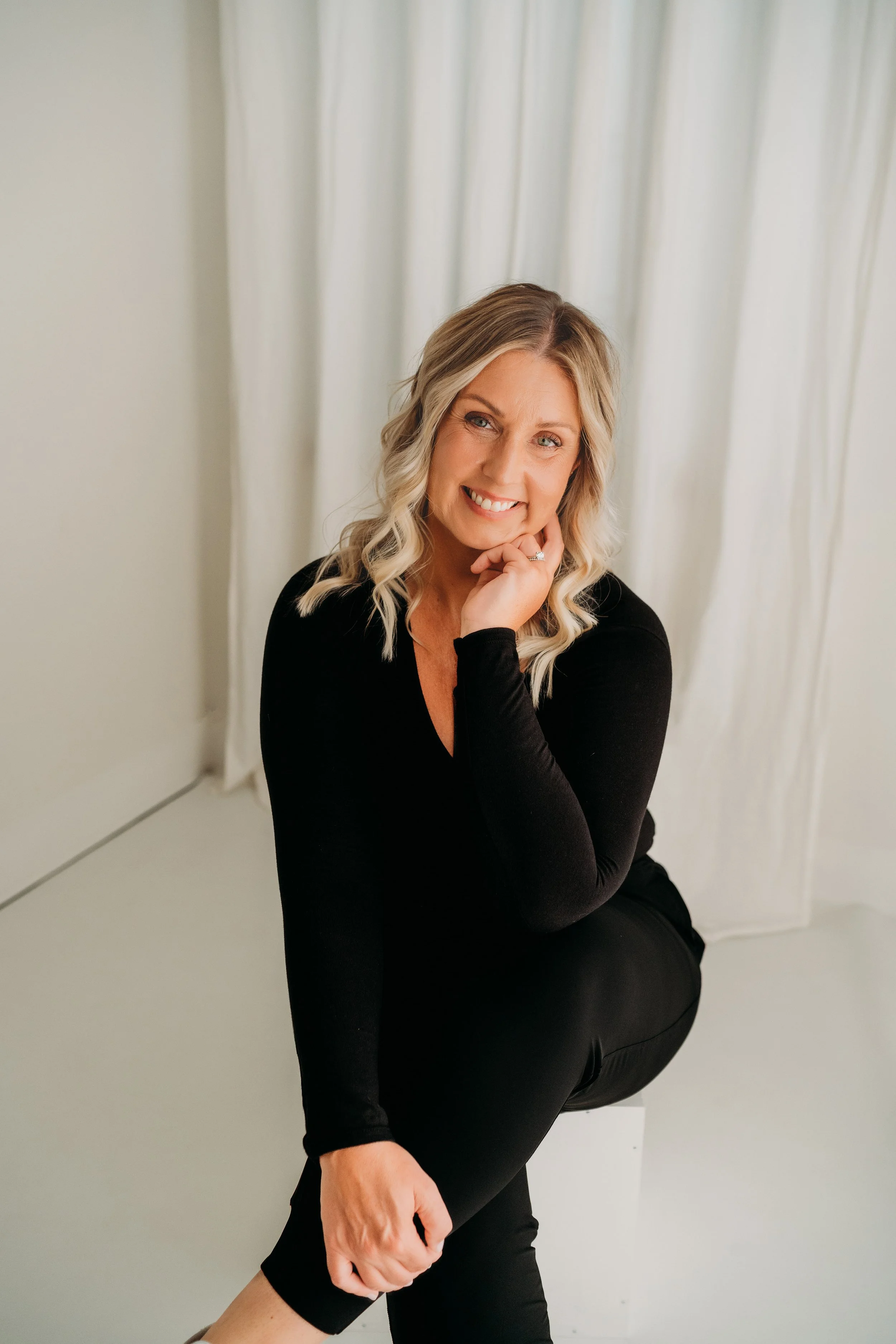 A woman with blonde hair in loose waves, wearing a black long-sleeve top, sitting on a white stool, smiling, with white curtains in the background.